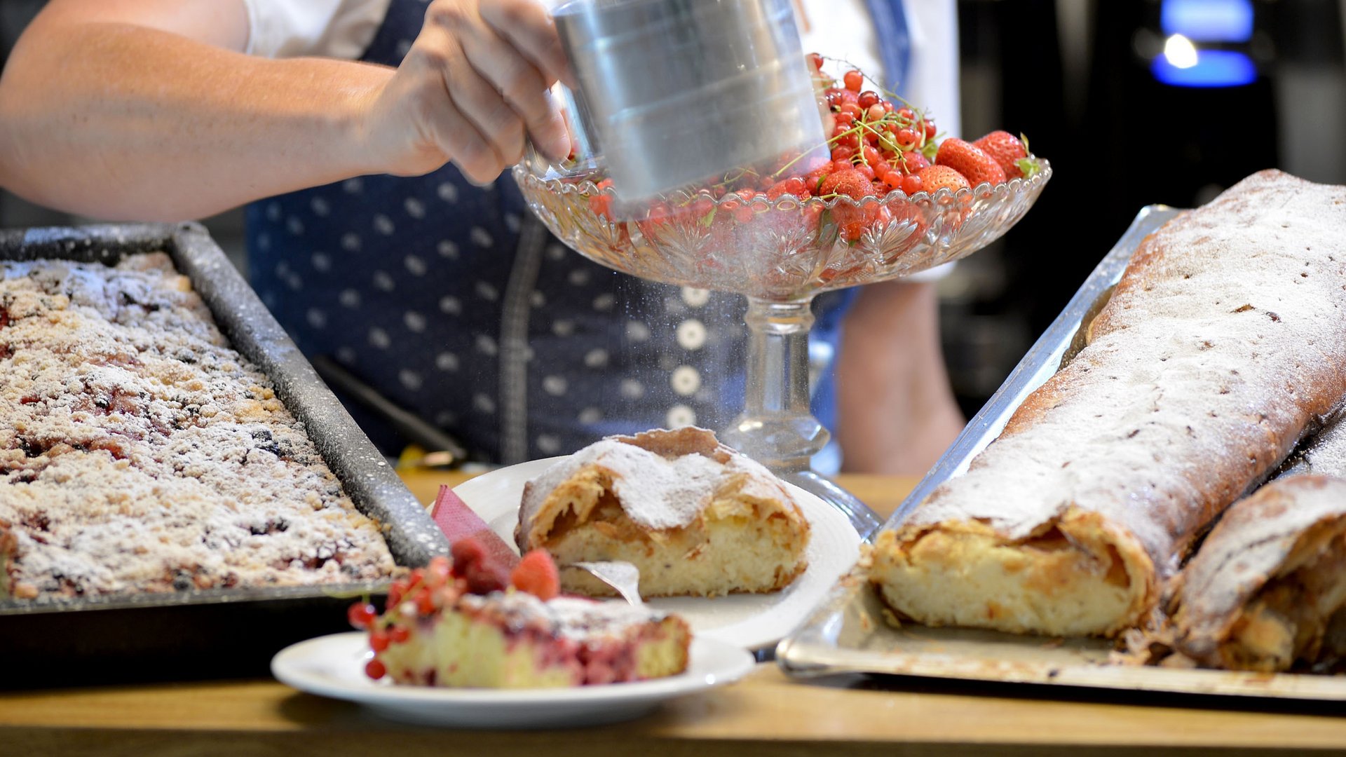 Person bestäubt Kuchen mit Puderzucker neben frischen Beeren