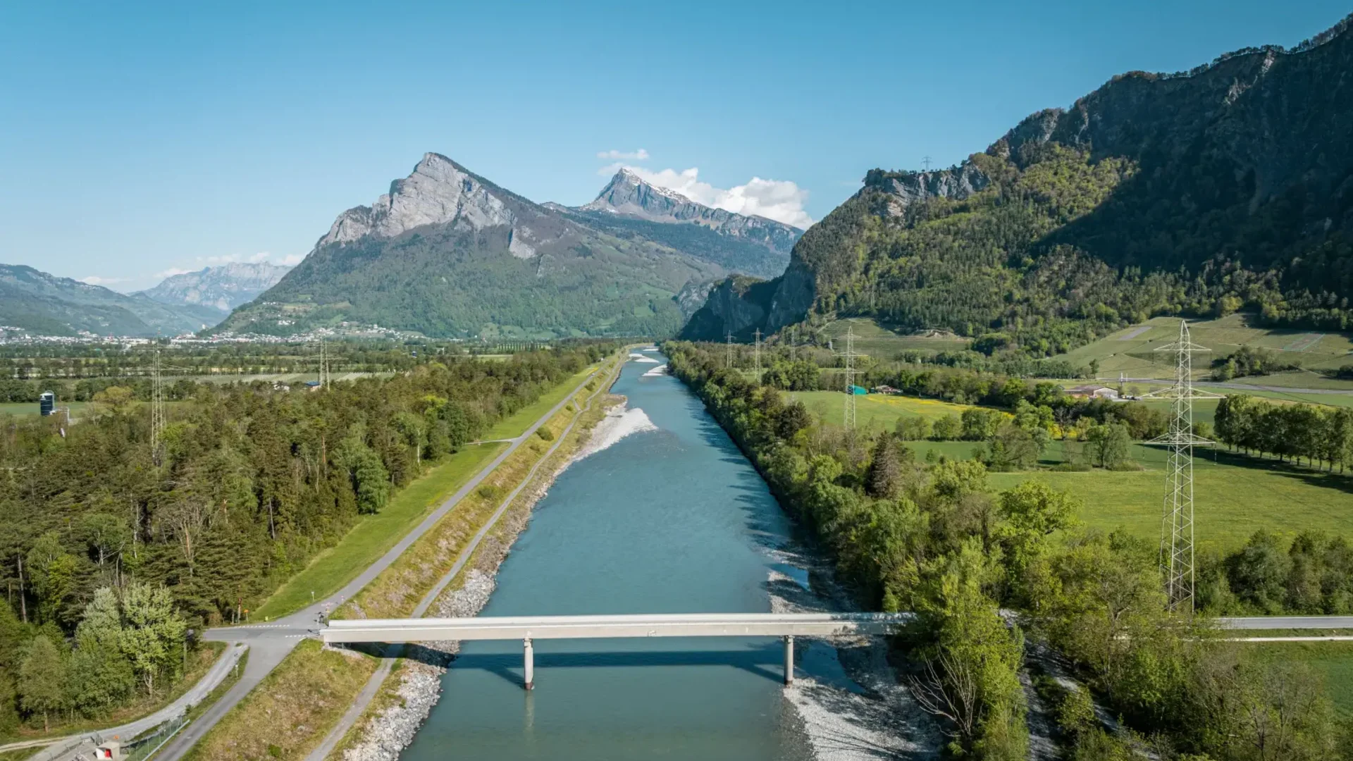 Fluss fließt durch Tal mit Bergen und Brücke bei klarem Himmel