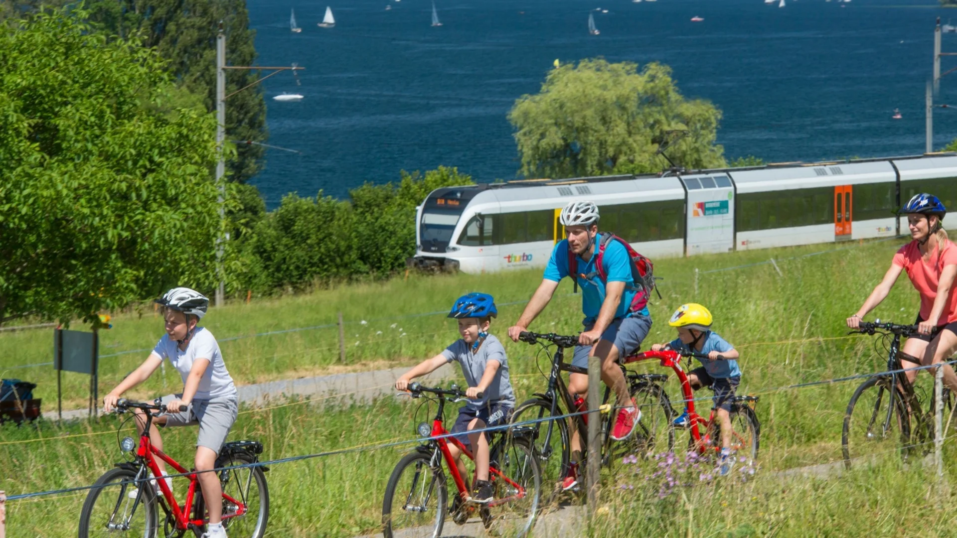 Family biking on a lakeside path with a train and sailboats in the background