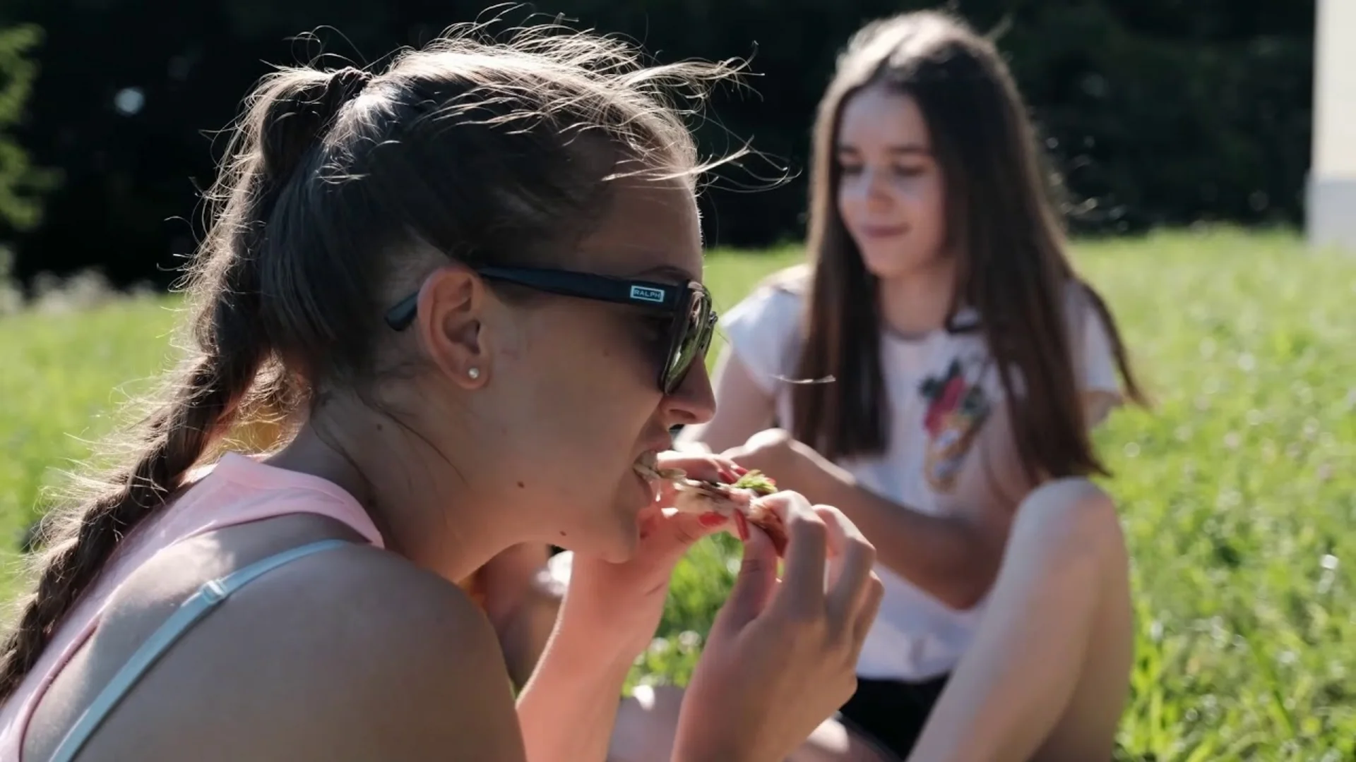 Twee meisjes zitten op gras, één eet een stuk pizza.