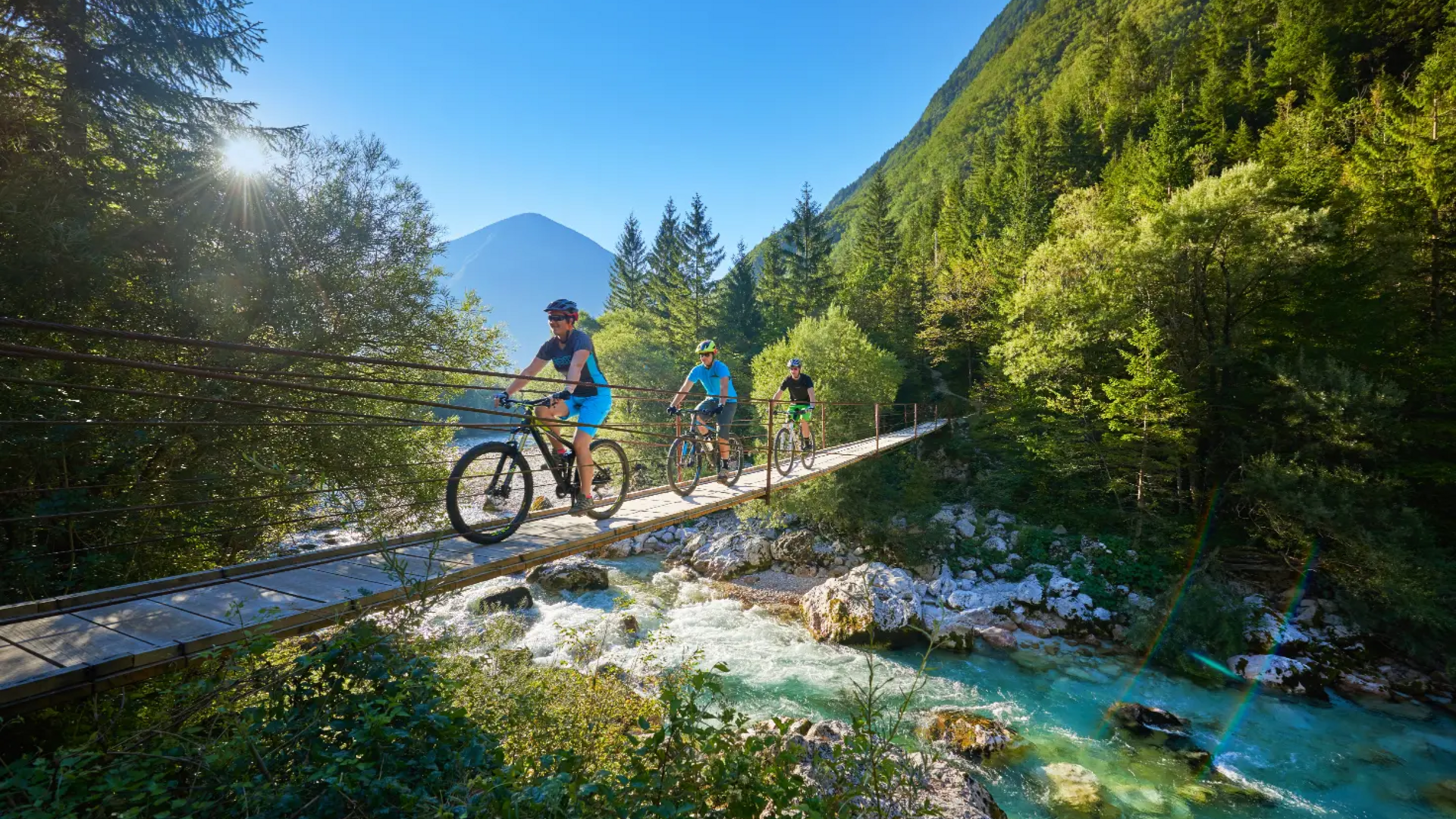 Three cyclists crossing suspension bridge over mountain river in sunny forest