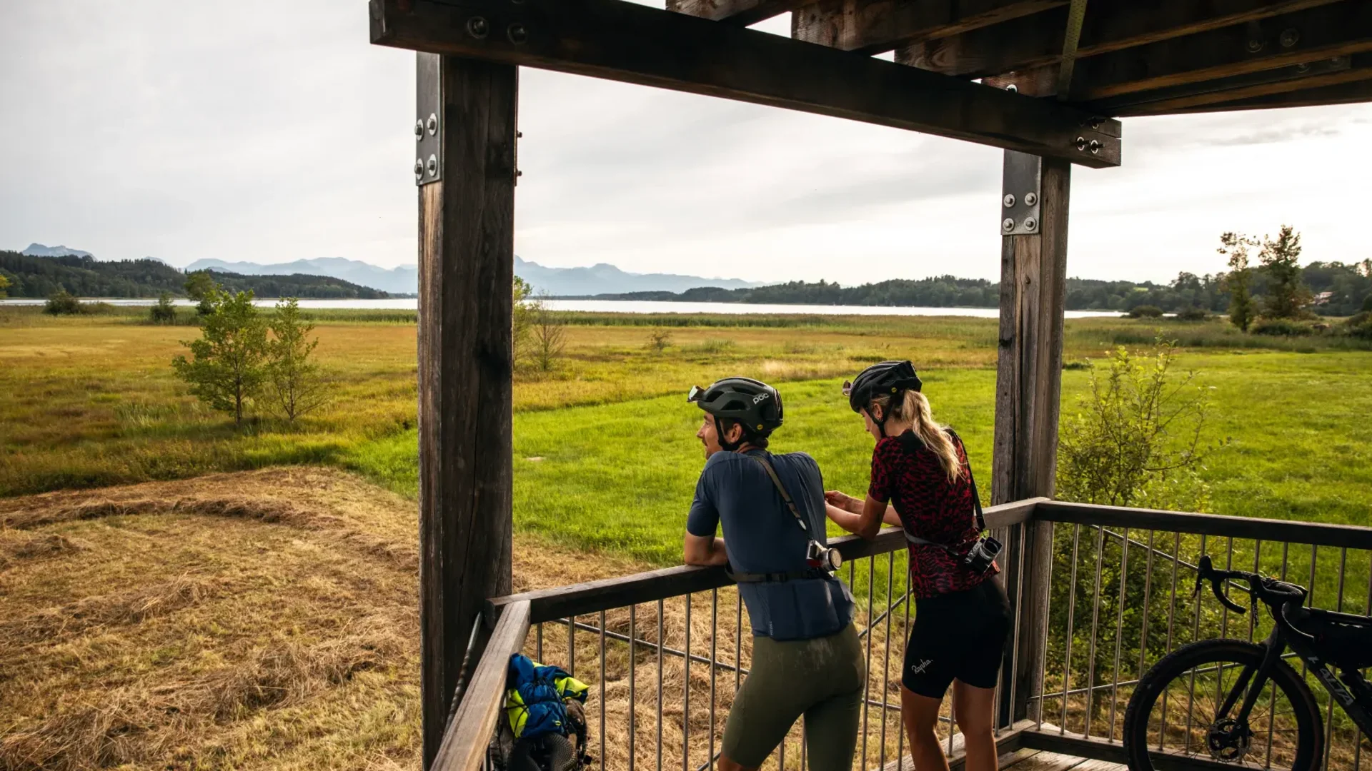 Zwei Radfahrer schauen von einem Holzaussichtsturm auf eine Landschaft mit See und Bergen