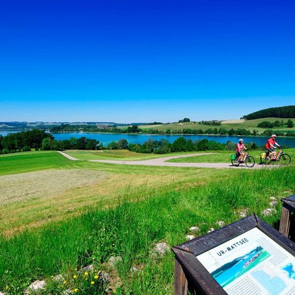 Tannberg Radrunde © Salzburger Seenland - Norbert Eisele-Hein Zwei Radfahrer auf einem Weg mit Blick auf einen See und Info-Tafeln auf einer grünen Wiese