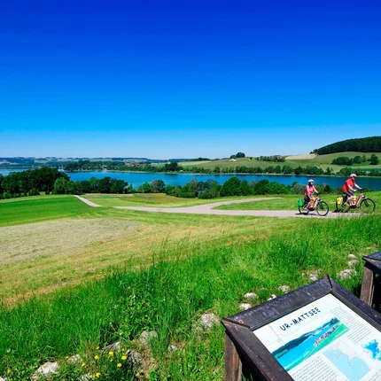 Twee fietsers op een pad met uitzicht op een meer en informatieborden op een groen weiland