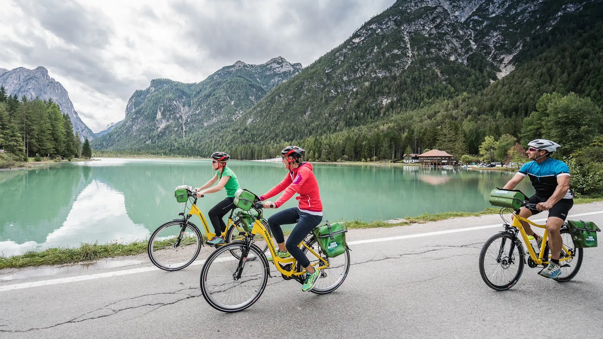 Three cyclists ride beside a lake with mountains in the background under cloudy sky