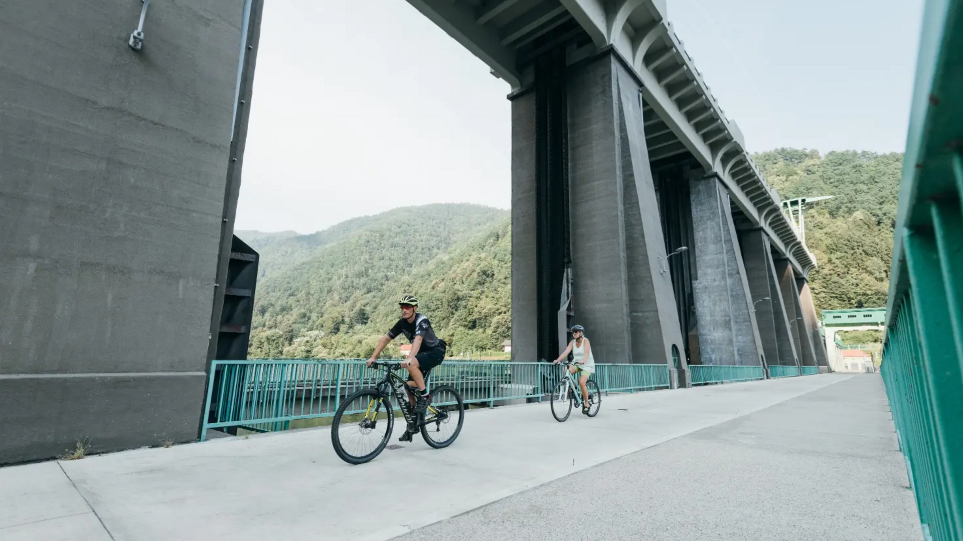 Two cyclists riding under a bridge in a mountainous area