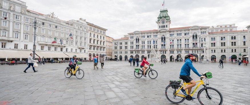 People cycling on a large square with historic buildings under a cloudy sky