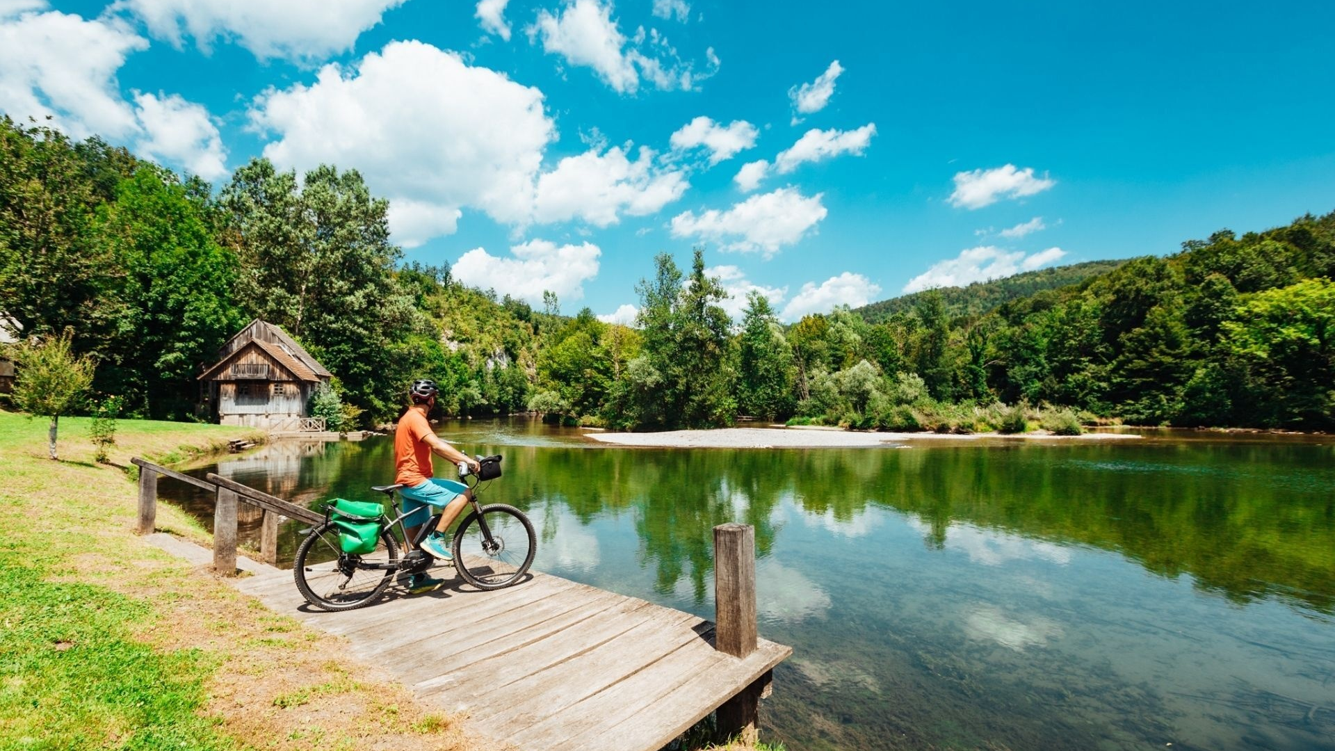 Cyclist on wooden pier by river with forest and blue sky