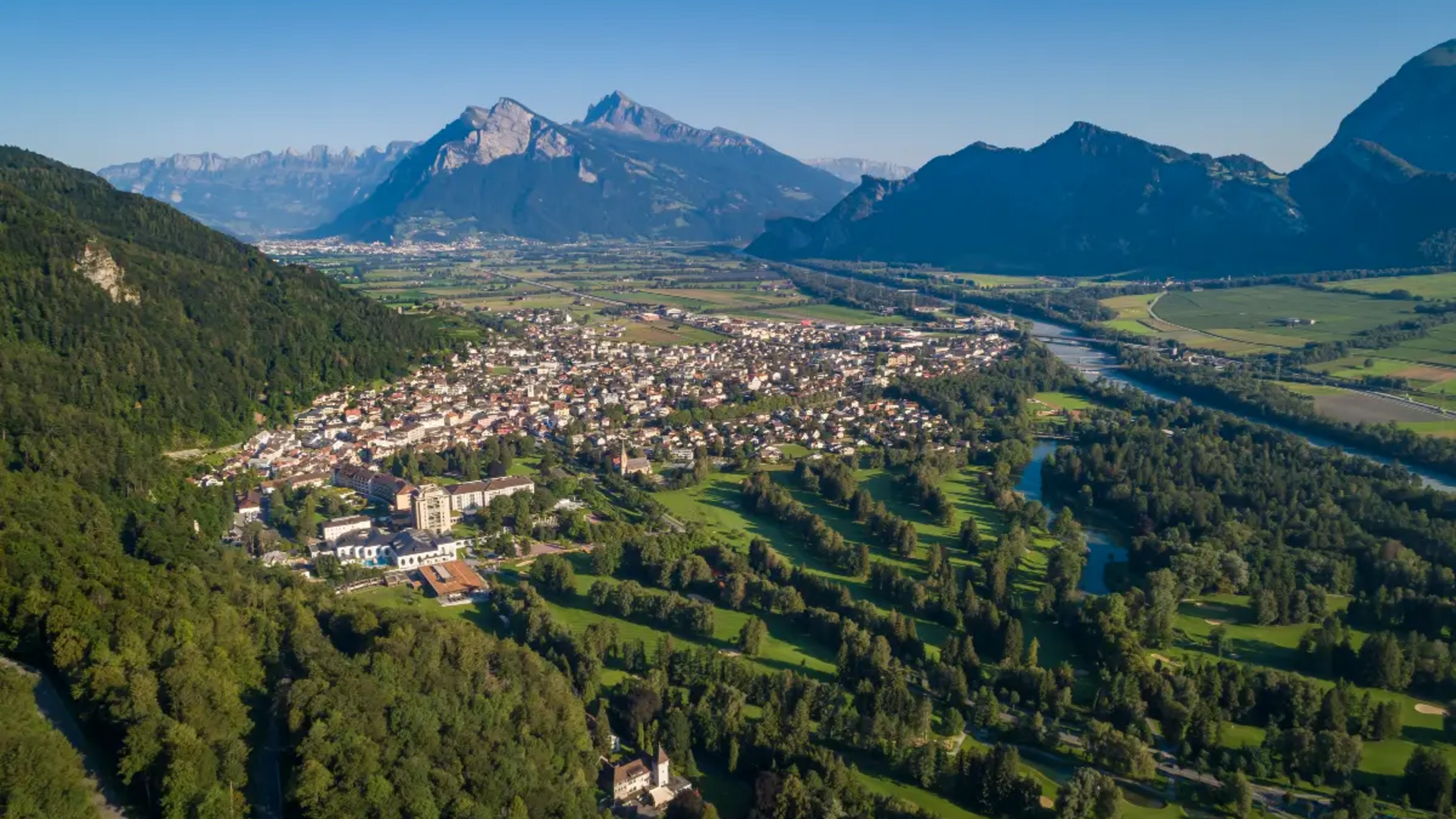 Luftaufnahme einer Stadt in einem grünen Tal mit Bergen und Fluss im Hintergrund
