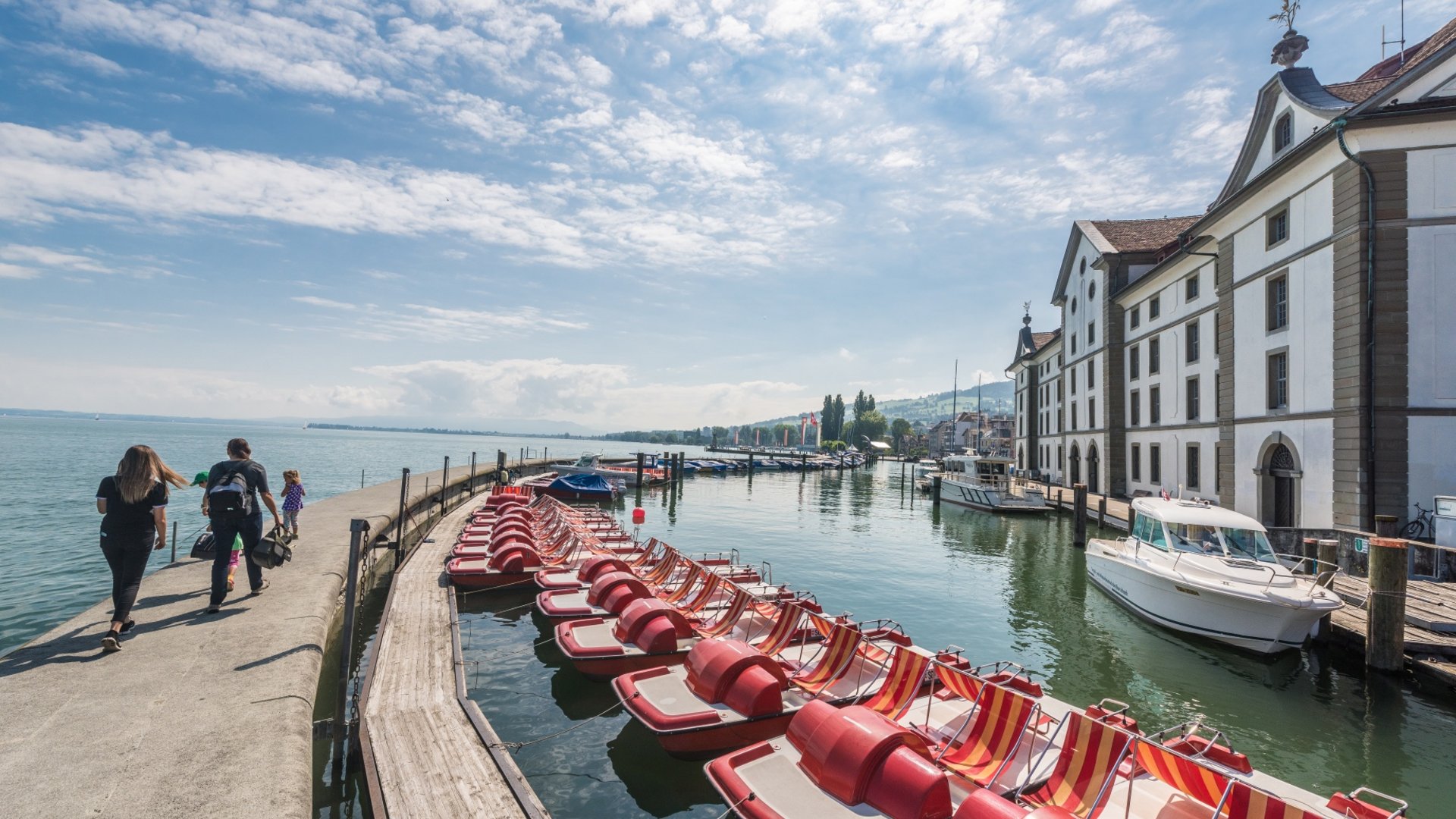 People walking near docked red pedal boats and historic buildings under blue sky