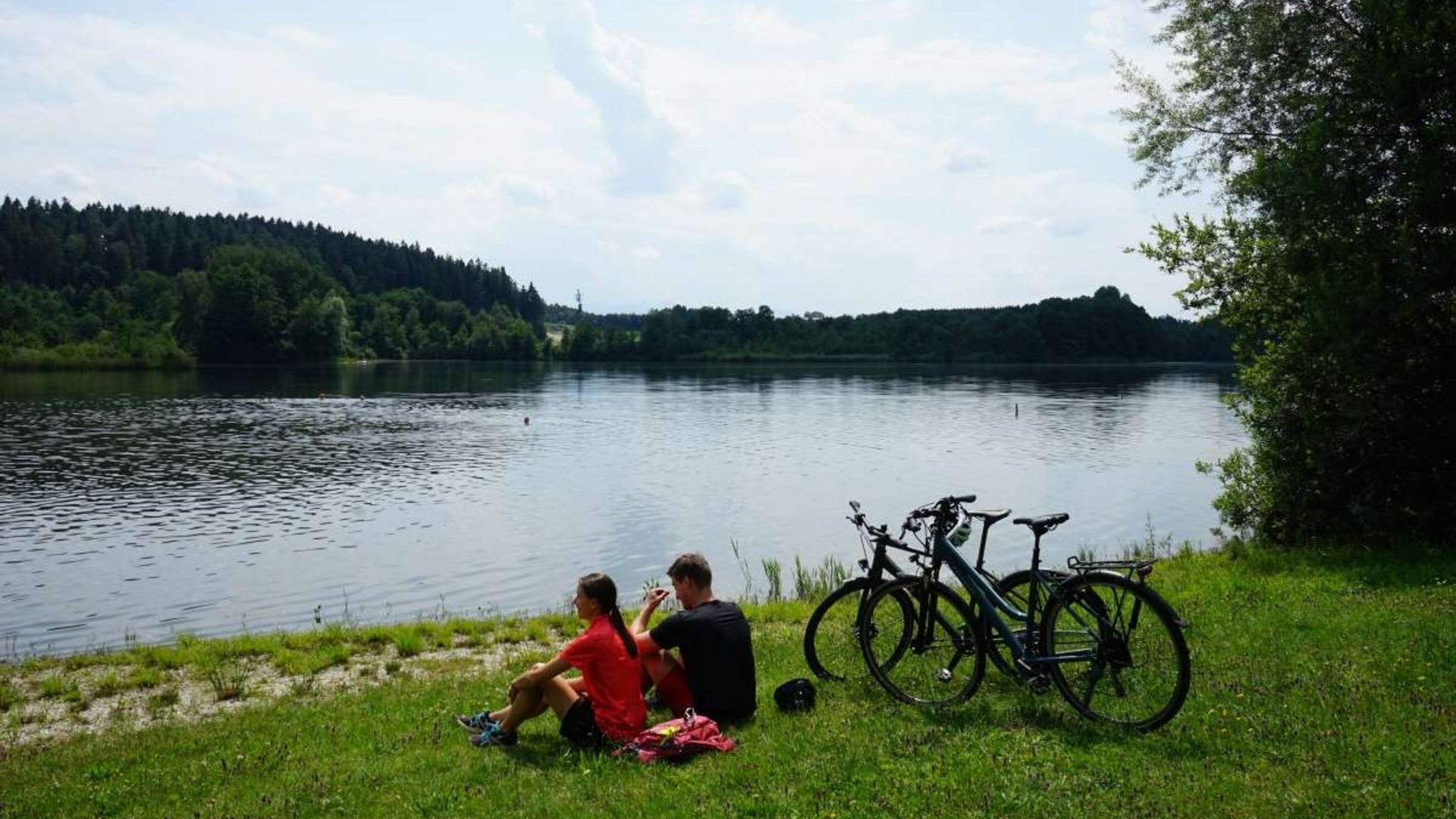 Two people sitting by the lake with bicycles on a grassy shore