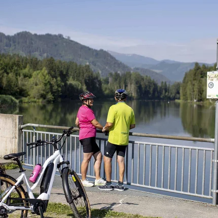 Two cyclists stop and look at a calm river with mountains in the background
