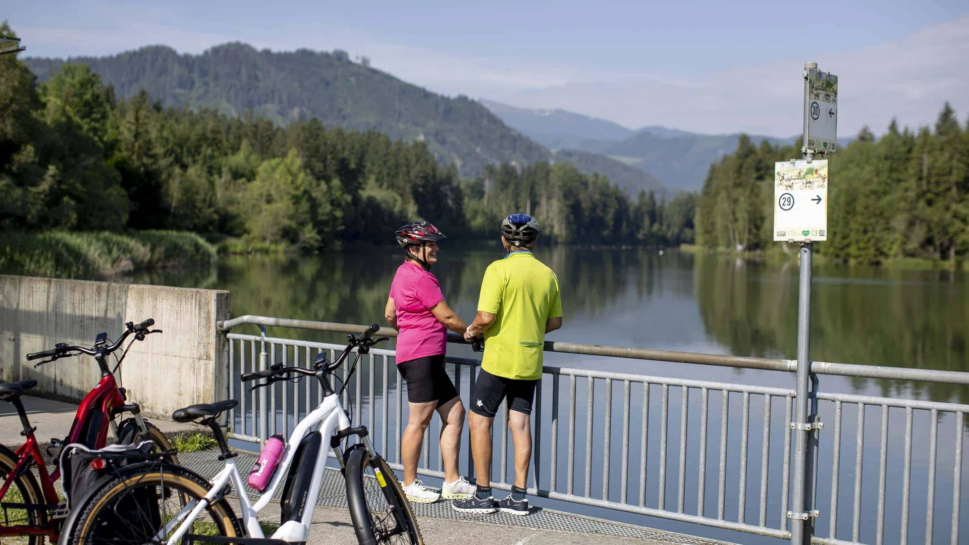 Twee fietsers stoppen en kijken naar een rustige rivier met bergen op de achtergrond