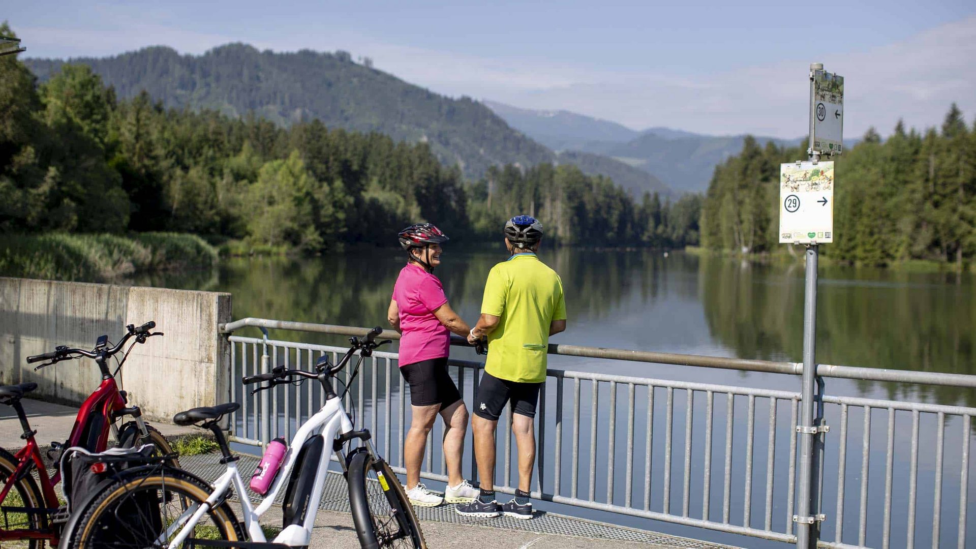 Two cyclists stop and look at a calm river with mountains in the background