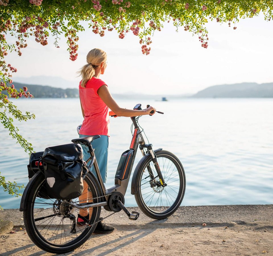 Packliste für einen perfekten Radurlaub © Kärnten Werbung - Gert Perauer Frau mit Fahrrad blickt auf ruhigen See und Berge bei Sonnenuntergang