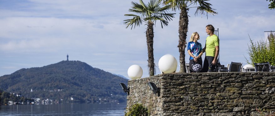 Couple holding bike helmets on stone wall by lake with mountain view