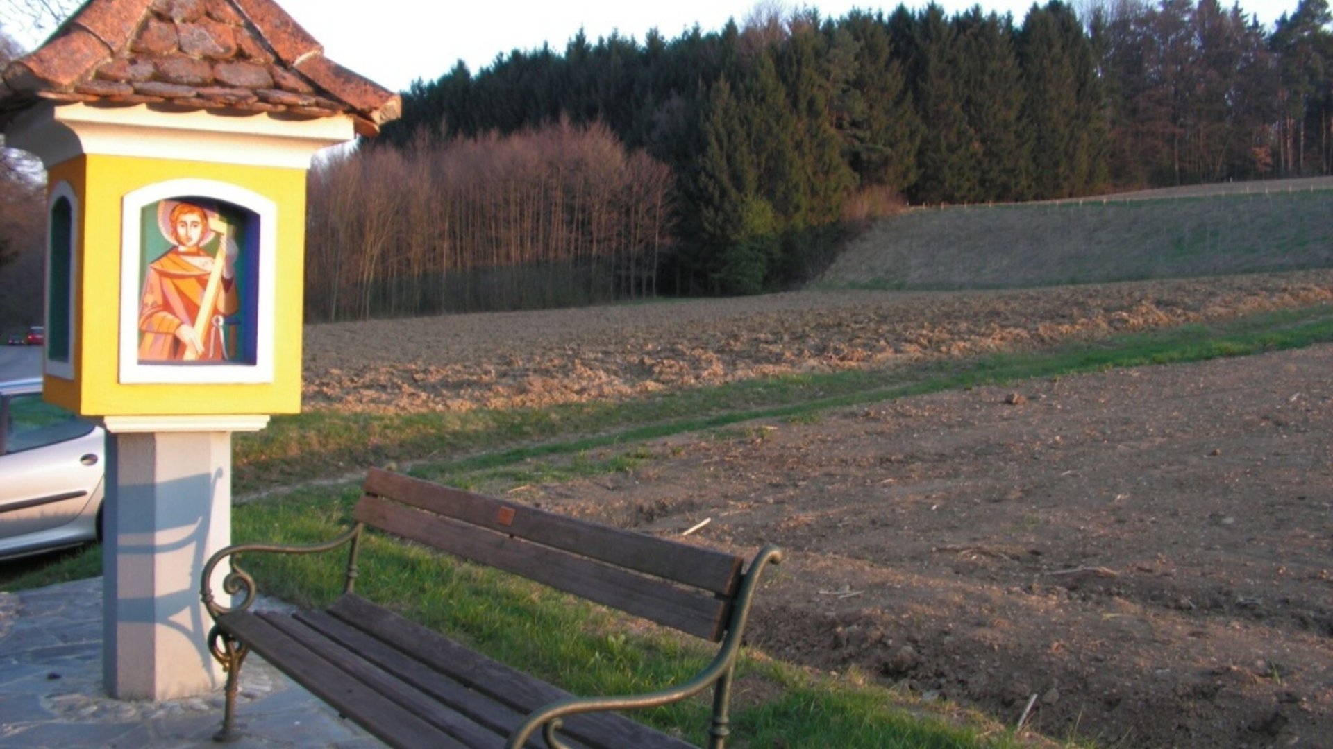 Wooden bench beside a religious wayside shrine near a field