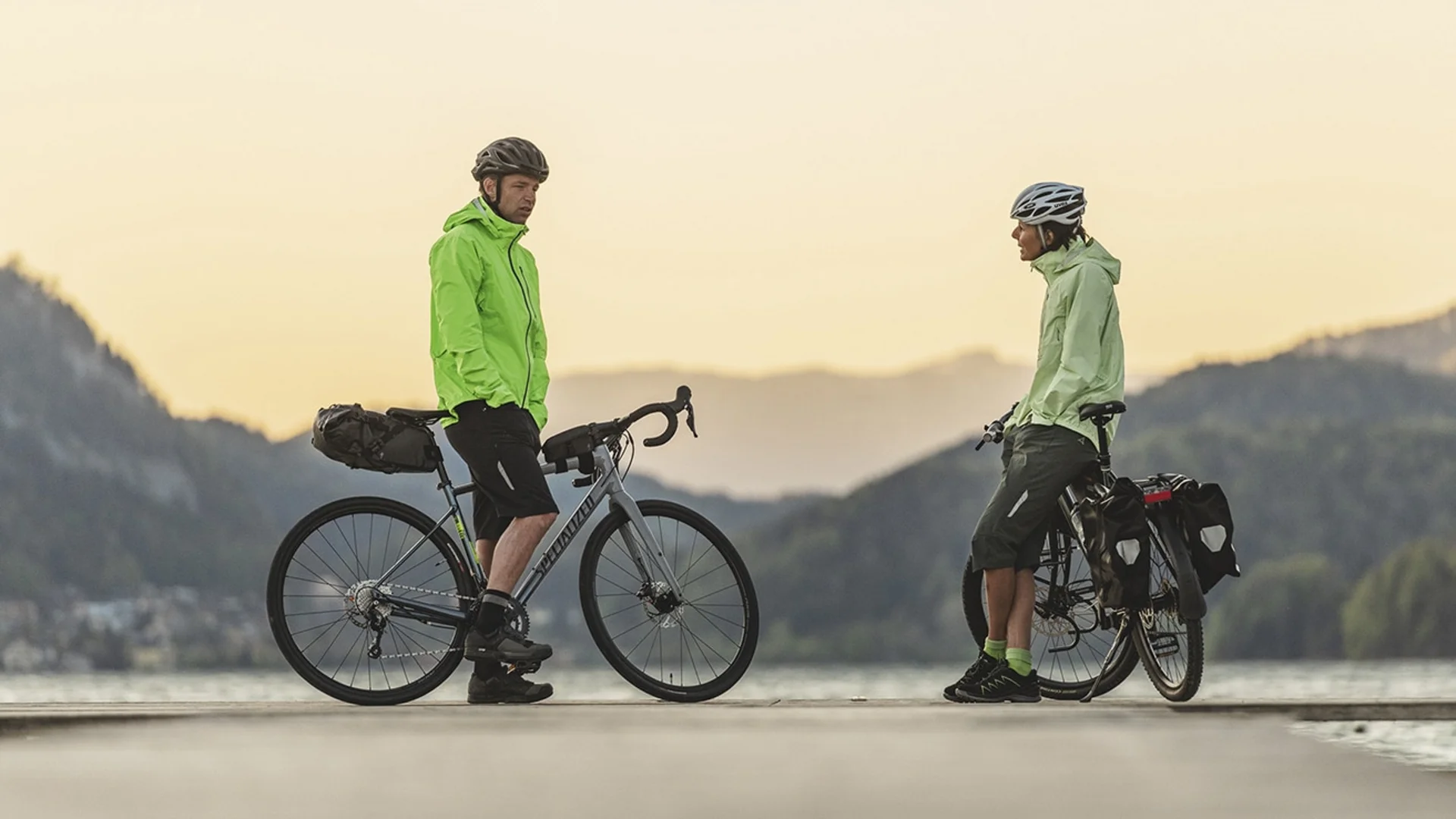 Two cyclists in green jackets standing with their bikes on a lakeside pier