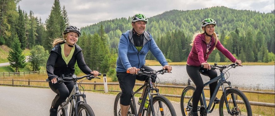 Three people cycling on a country road near forest and lake