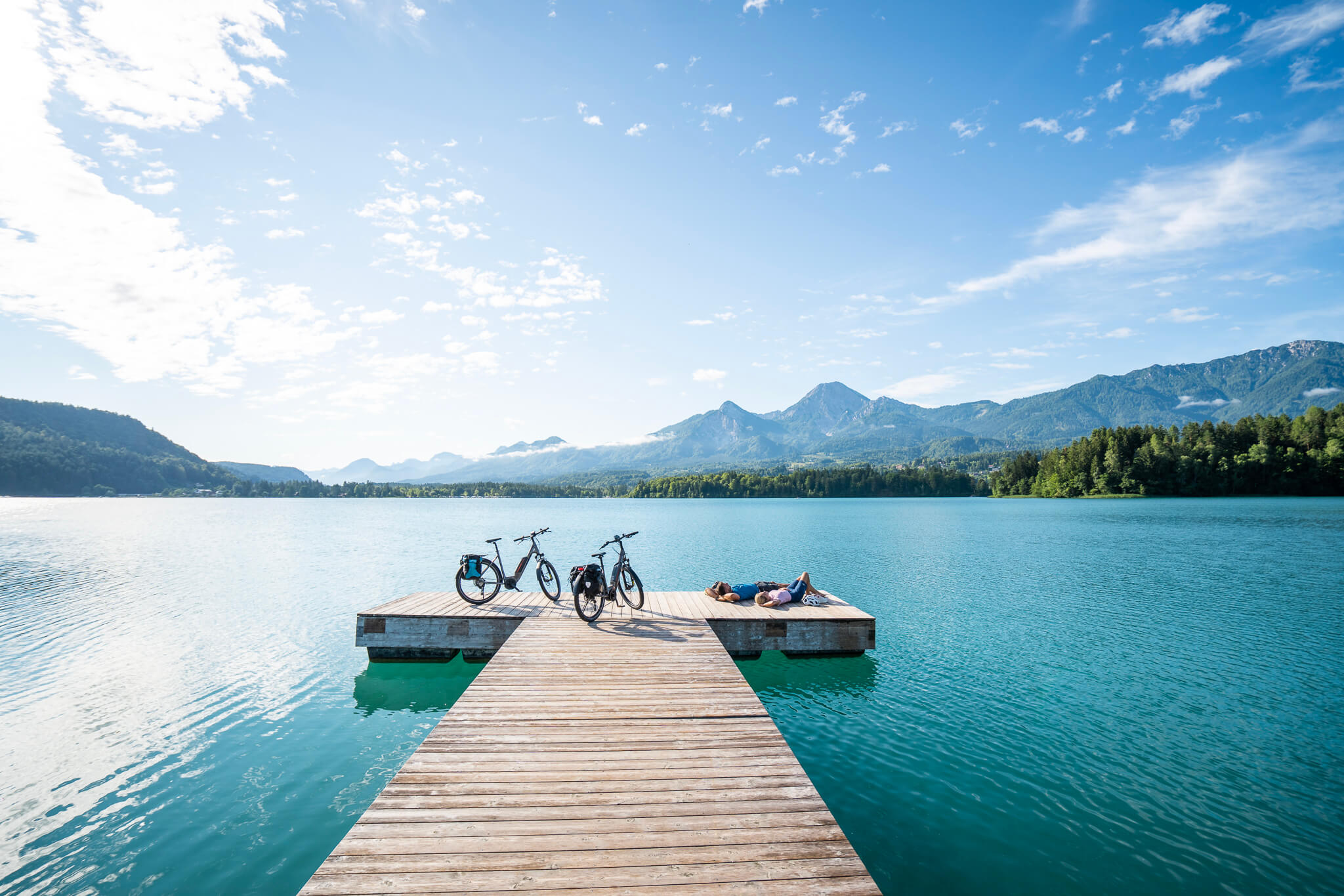 Steg mit Fahrrädern und zwei Menschen am klaren See vor Bergen