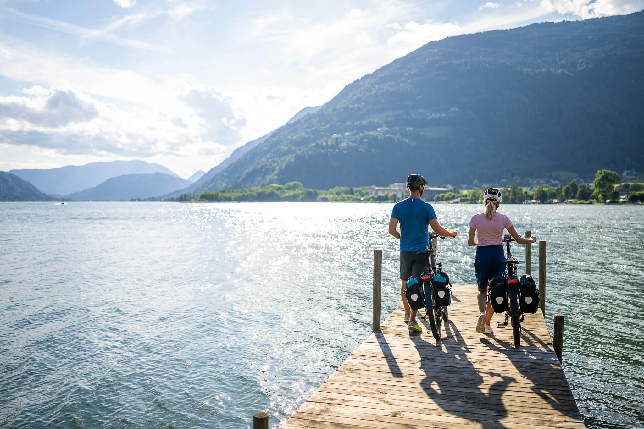 Zwei Radfahrer mit Helmen auf einem Steg am See mit Bergblick