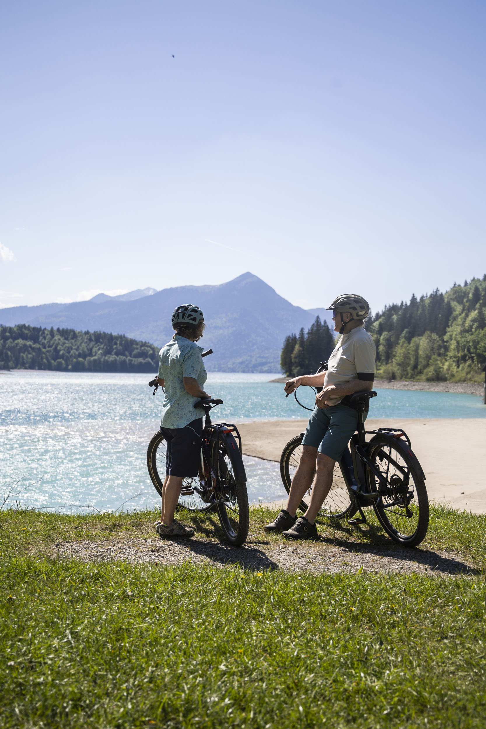 Zwei Radfahrer mit Helm machen Pause an einem See mit Bergen im Hintergrund