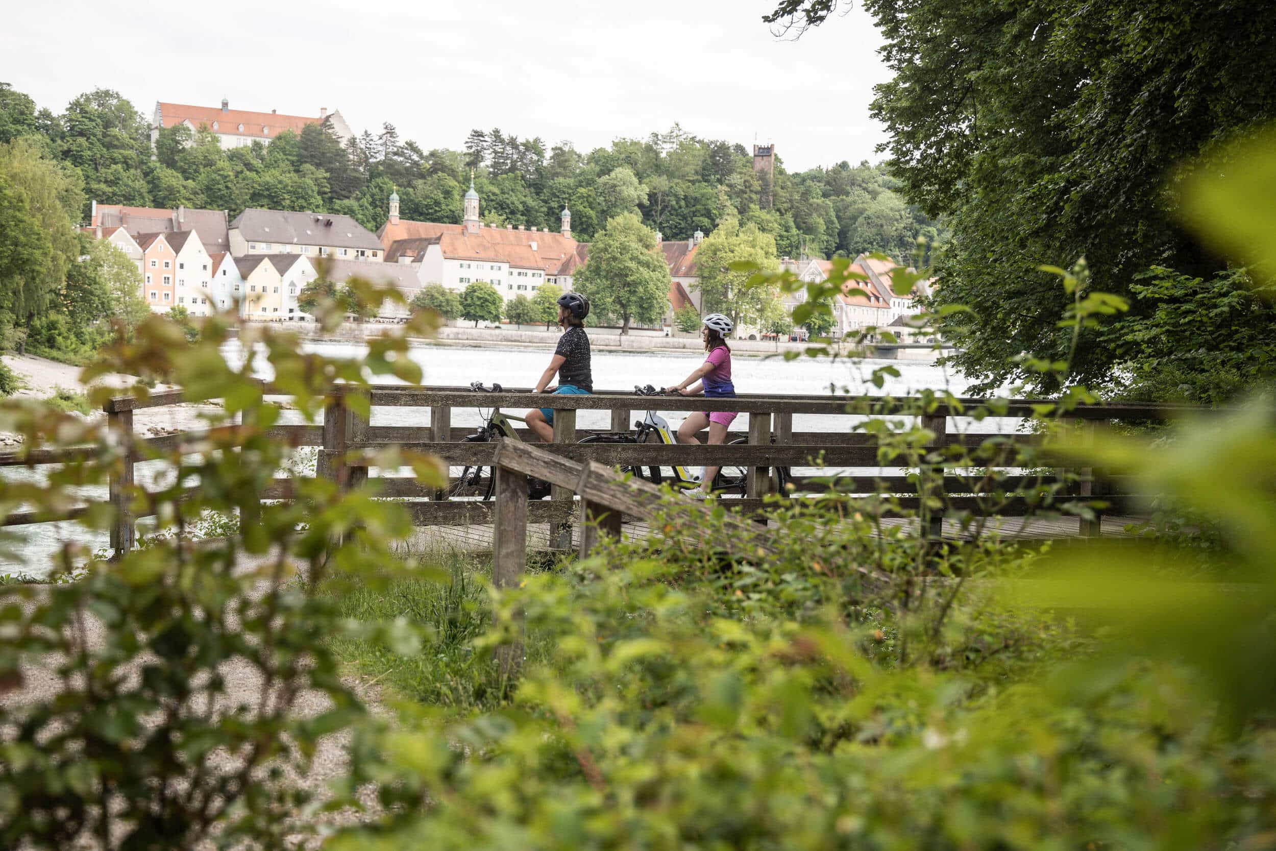 Zwei Radfahrer auf einem Holzsteg mit Blick auf einen Fluss und Häuser