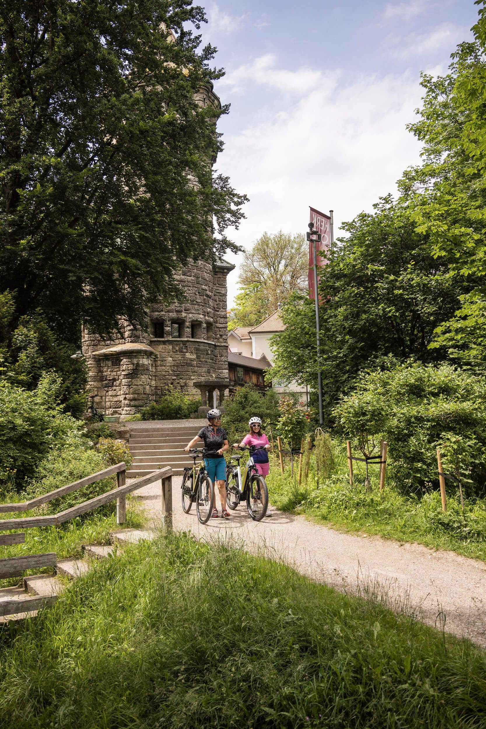 Zwei Radfahrerinnen auf einem Weg vor einem alten steinernen Turm im Grünen