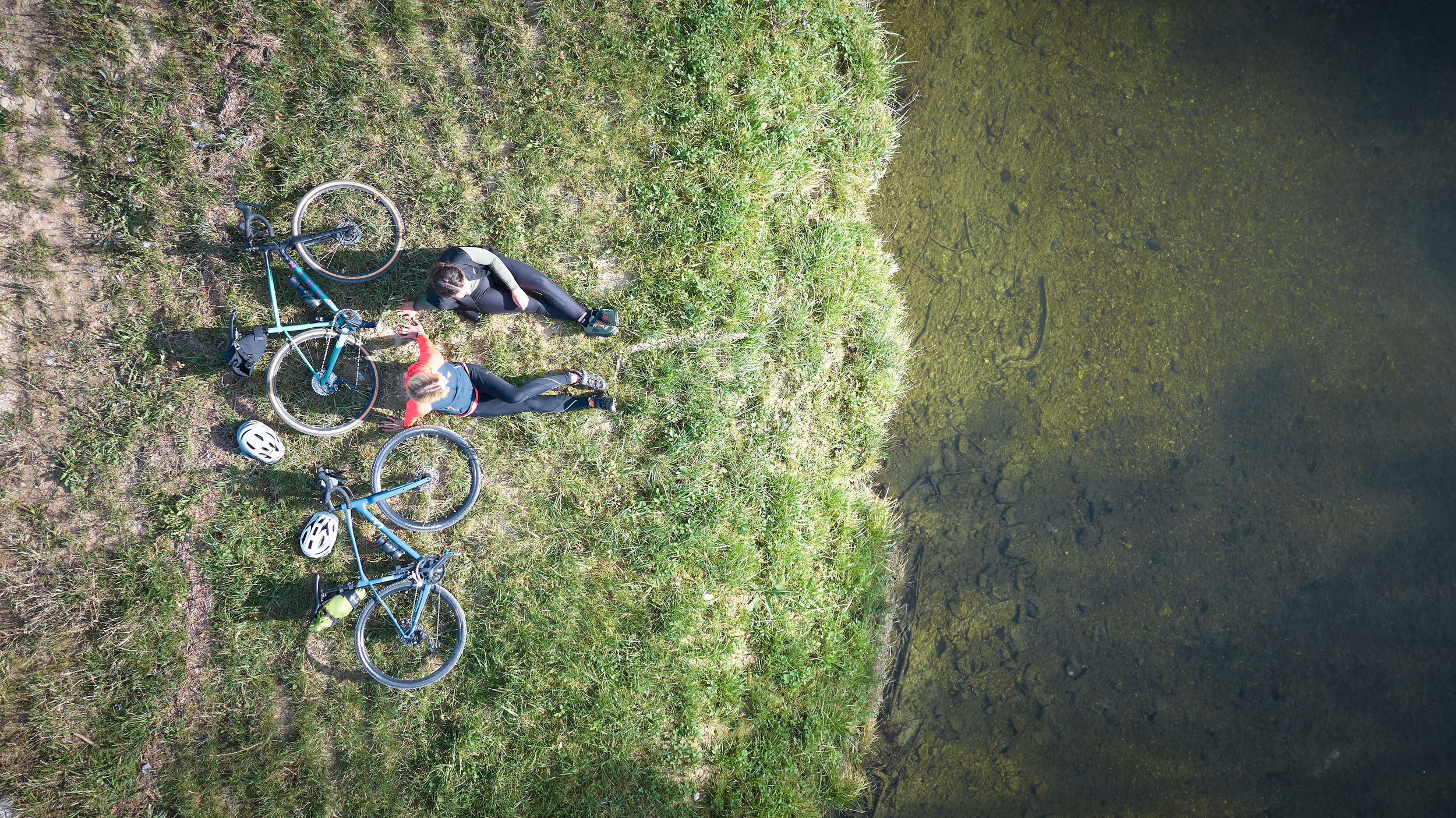 Zwei Radfahrer sitzen auf Gras am Ufer eines Gewässers mit ihren Fahrrädern