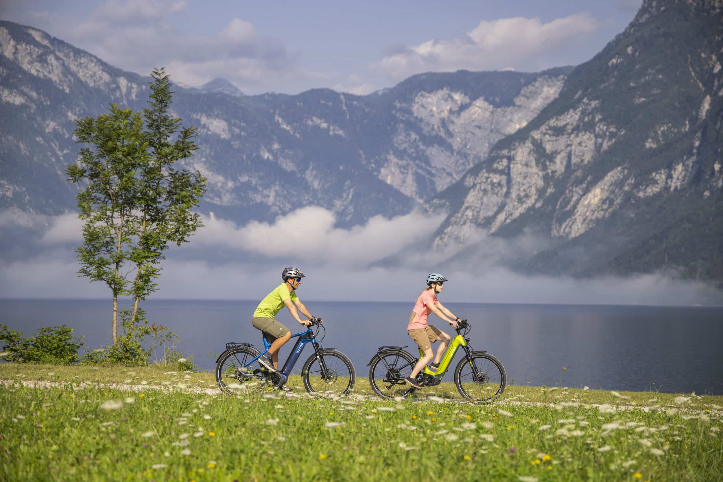 Zwei Radfahrer mit Helmen fahren an einem See mit Bergen im Hintergrund