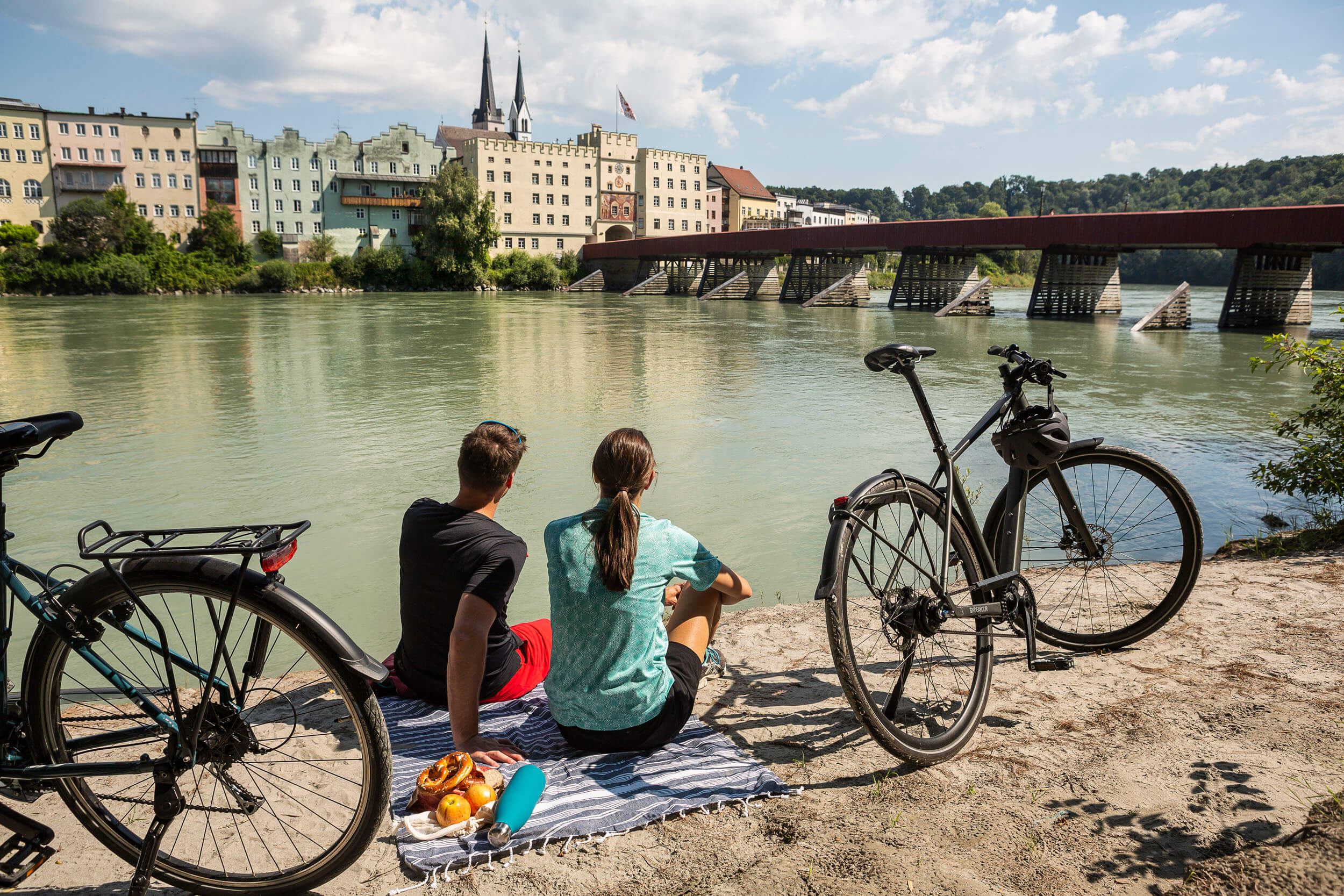 Zwei Personen sitzen am Fluss mit Fahrrädern und Blick auf historische Gebäude