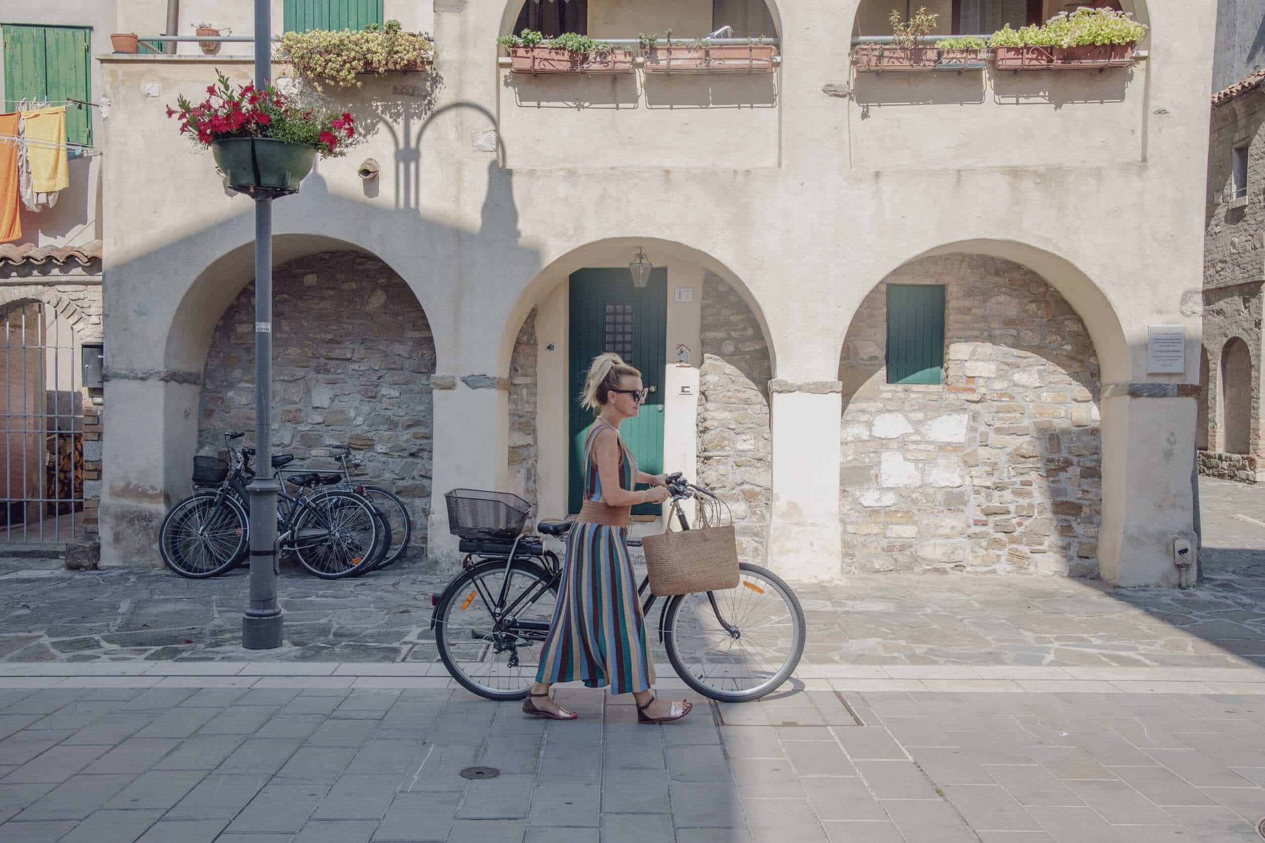 Frau schiebt Fahrrad auf gepflastertem Weg vor Gebäude mit Bögen