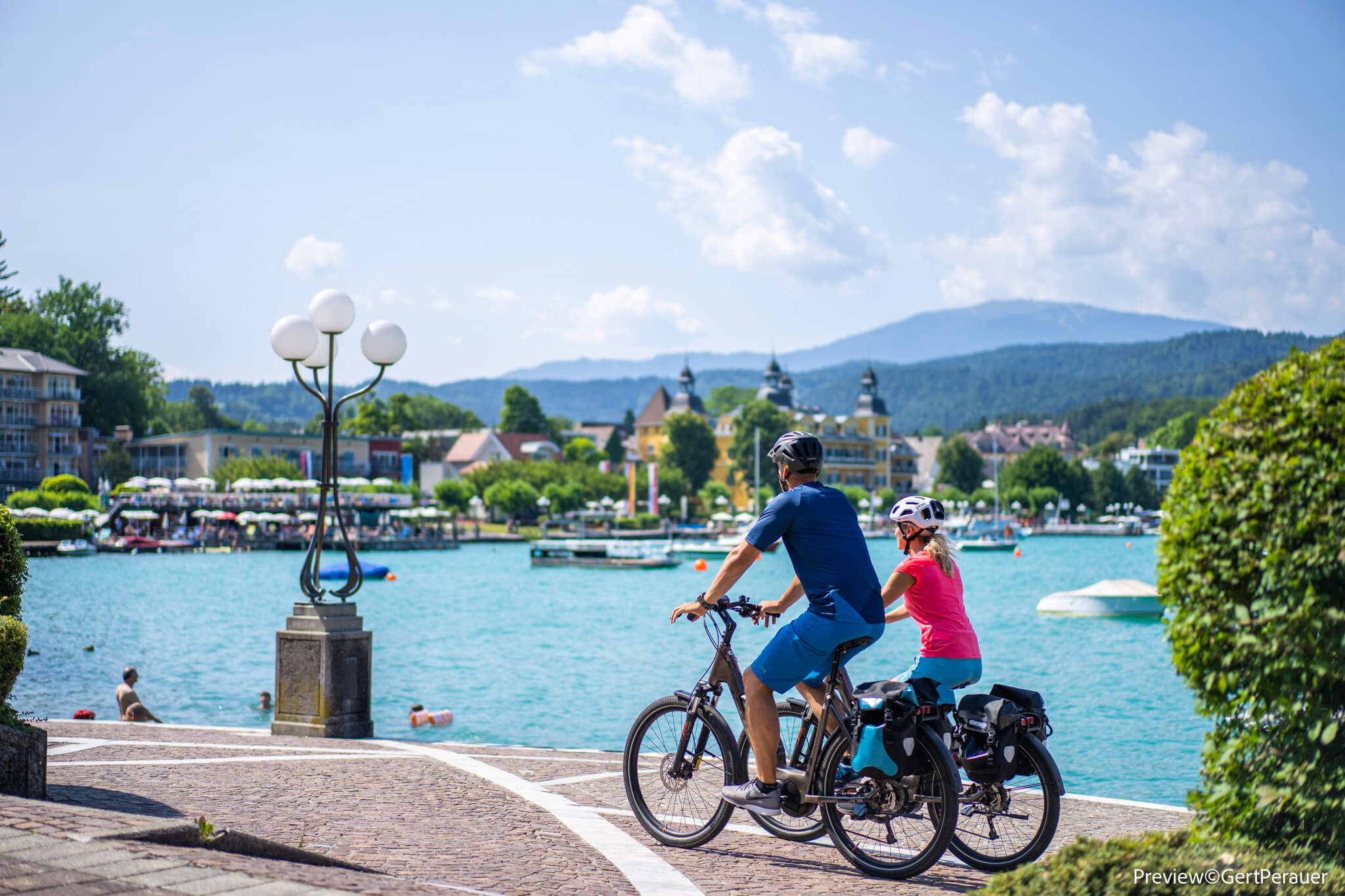 Zwei Radfahrer am Ufer eines Sees mit Bergen und Häusern im Hintergrund