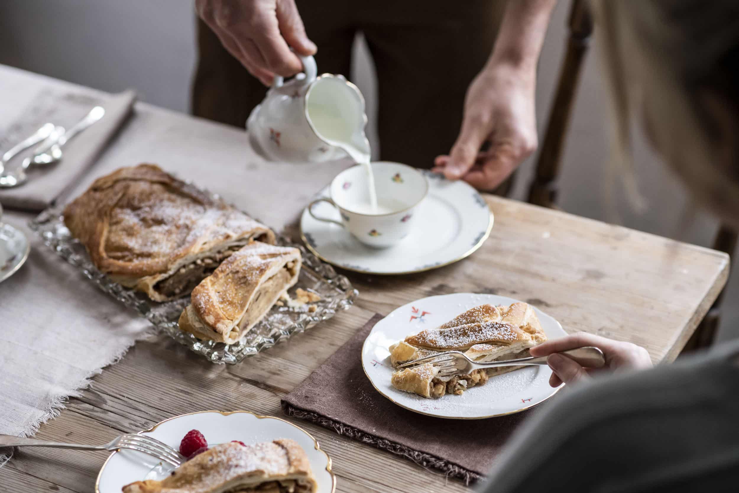 Person pouring milk into cup with apple strudel on wooden table