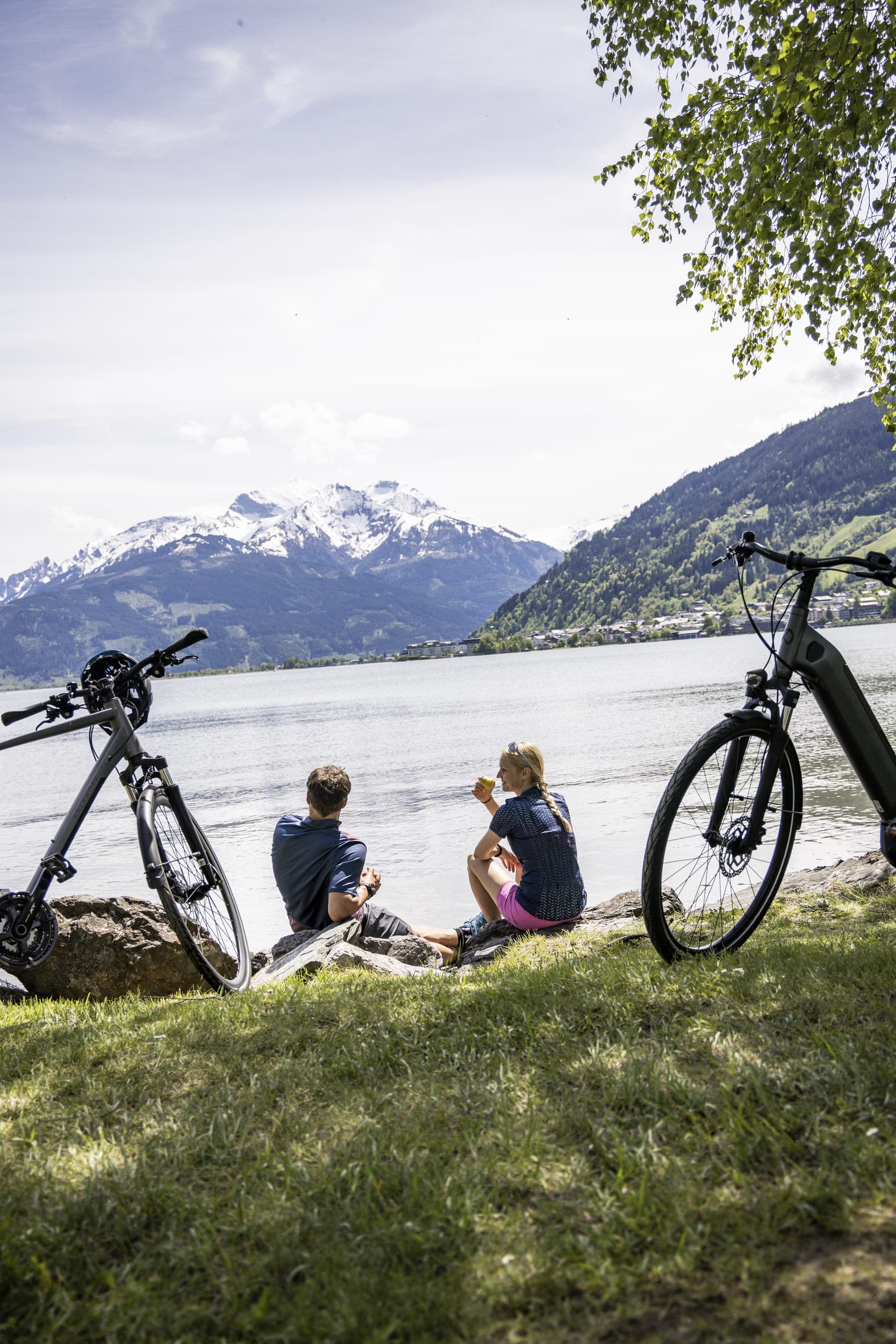Pärchen sitzt am Seeufer mit Fahrrädern und Blick auf verschneite Berge