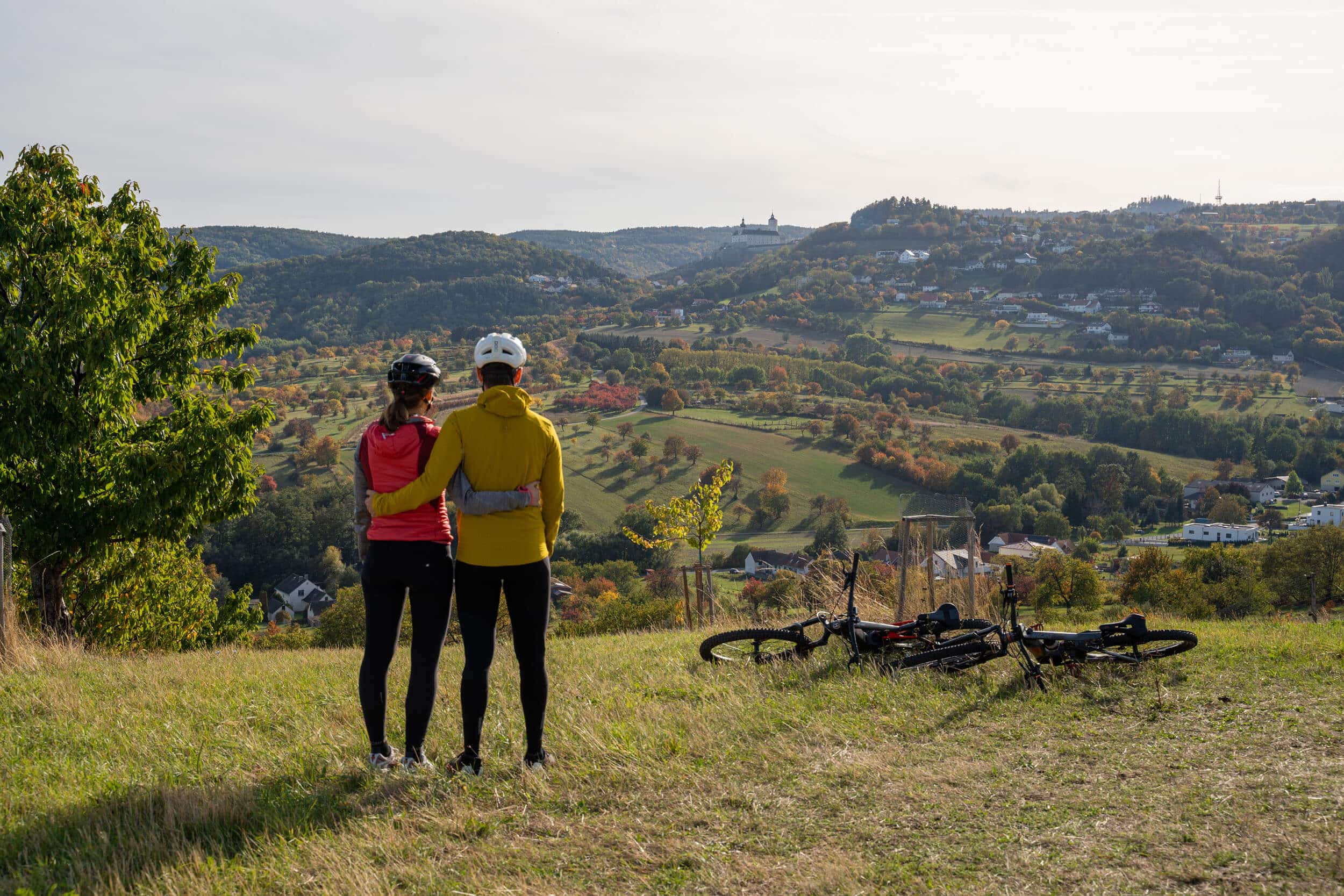 Paar mit Fahrradhelmen schaut auf eine hügelige Landschaft mit Gebäuden und Bäumen.