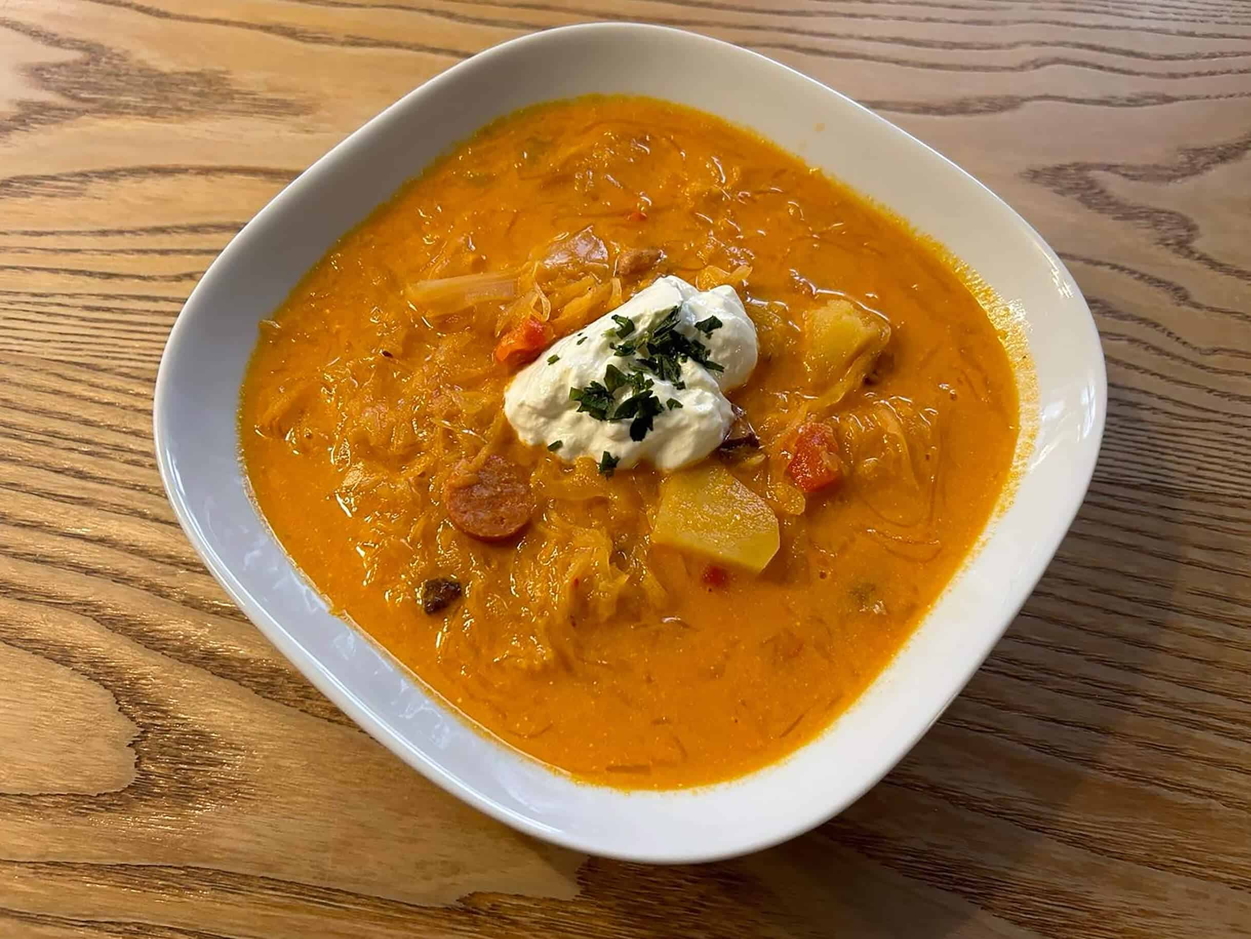 Bowl of sauerkraut soup with potatoes, sausage, and sour cream on wooden surface.