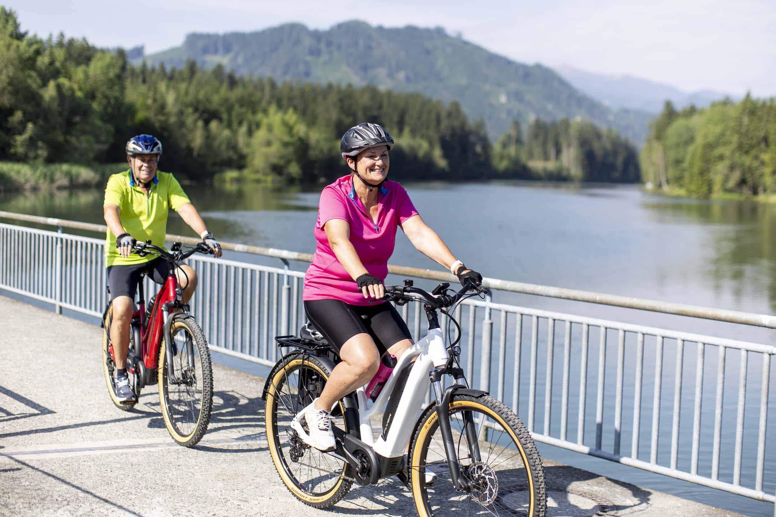 Zwei Senioren fahren Fahrräder auf einem Weg neben einem Fluss mit Bergen im Hintergrund