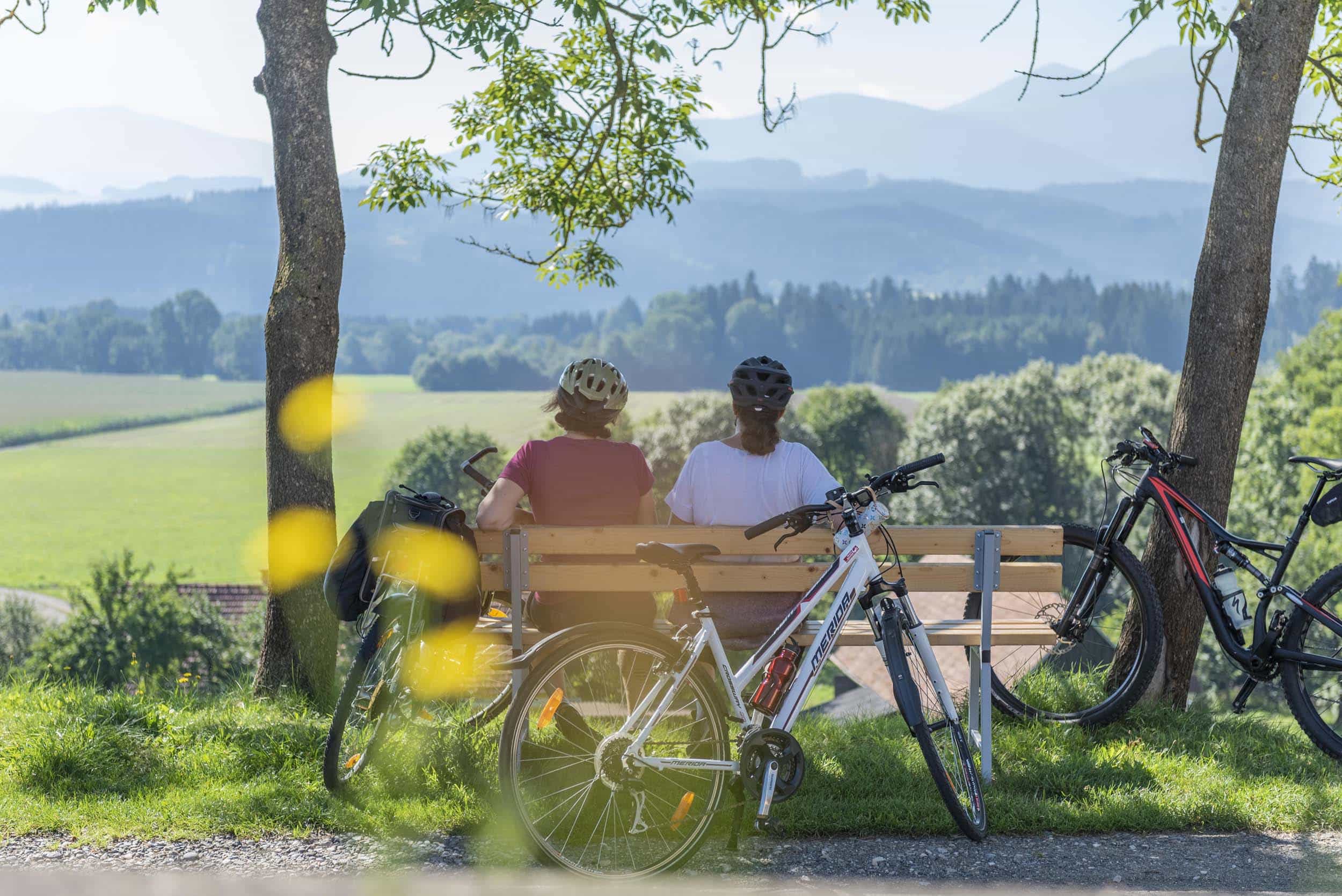 Zwei Radfahrer sitzen auf einer Bank mit Blick auf grüne Landschaft und Berge