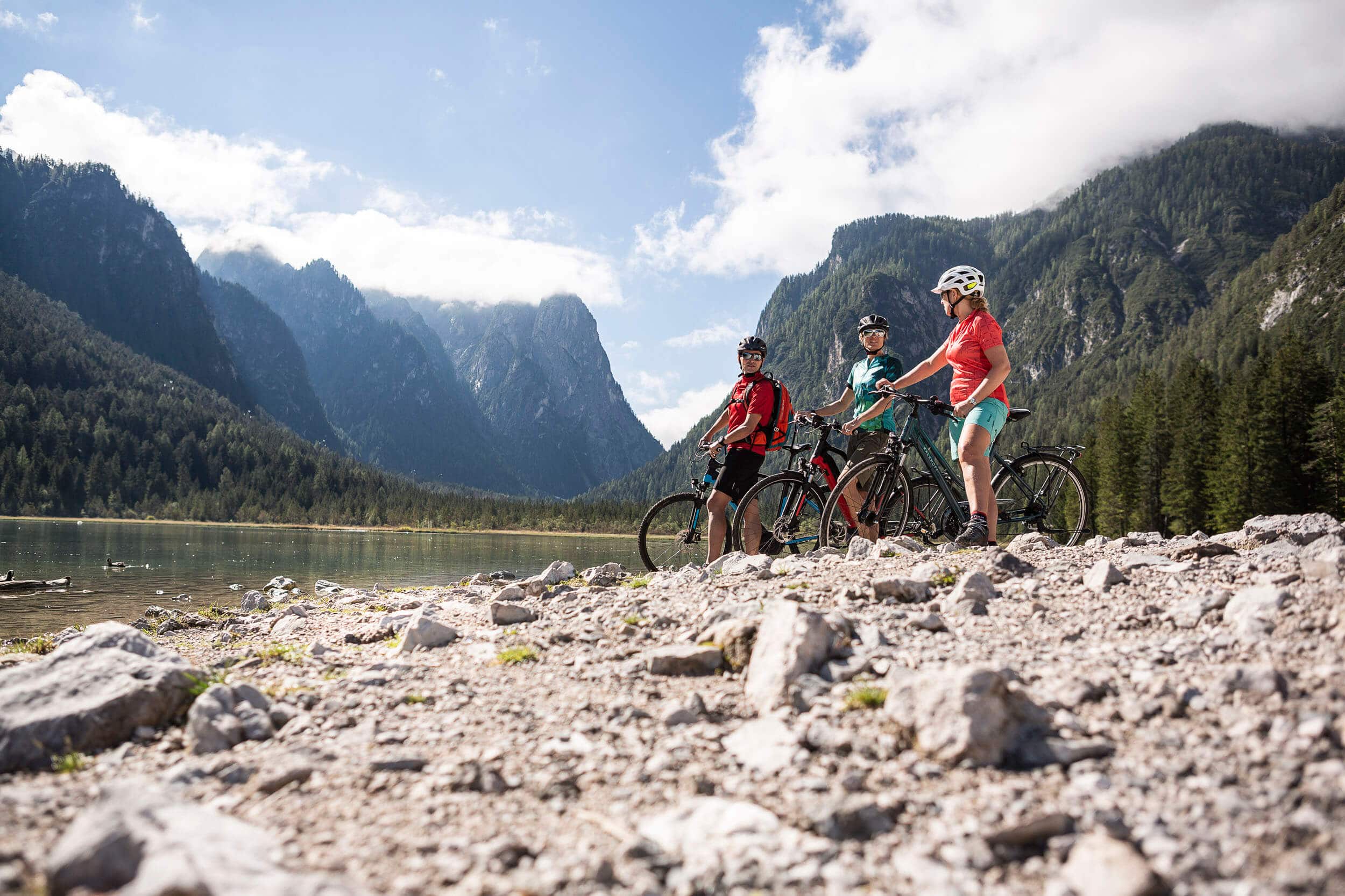 Drei Radfahrer stehen an einem Bergsee mit Alpen im Hintergrund