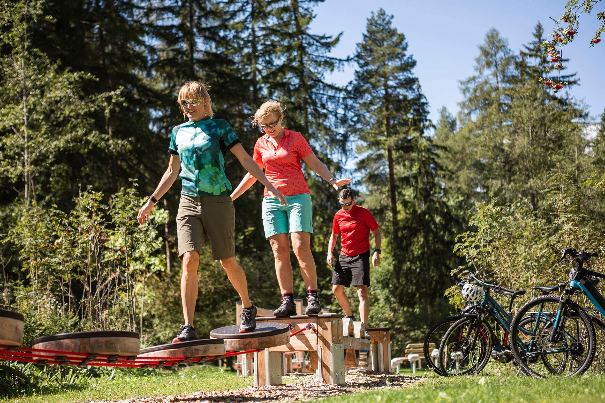 Drei Personen balancieren auf einem Holzparcours im Wald bei sonnigem Wetter
