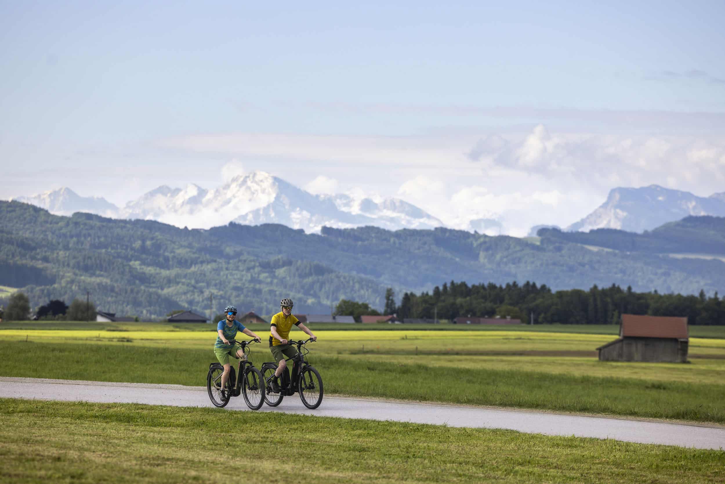 Zwei Radfahrer fahren auf einem Weg durch grüne Wiesen mit Bergen im Hintergrund