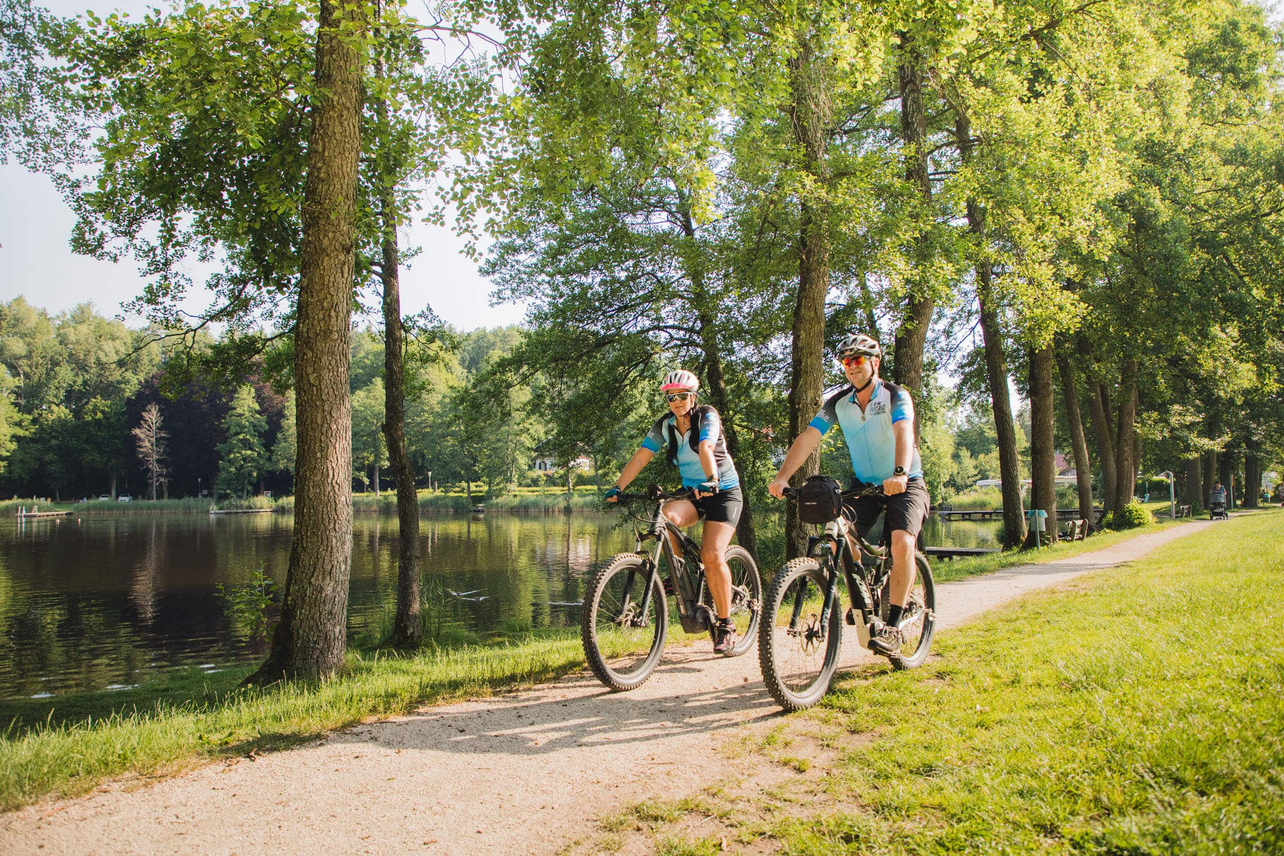 Zwei Radfahrer fahren auf einem Waldweg neben einem See bei Sonnenschein