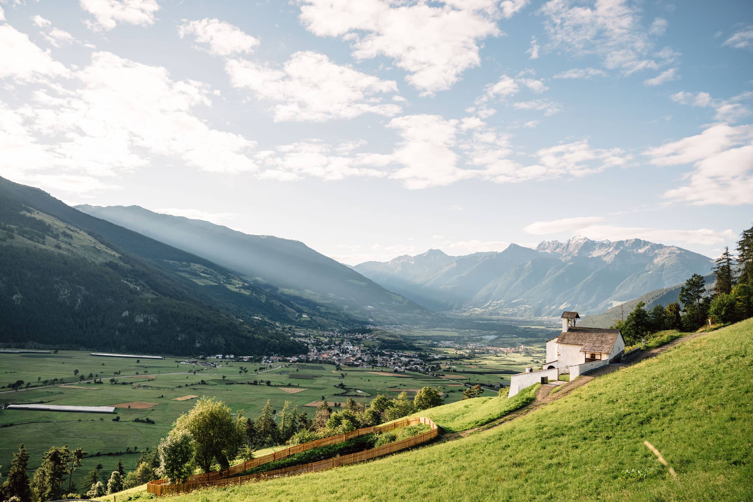 3 Highlights für den Radurlaub am Reschensee © Benjamin Pfitscher Bergtal mit Kirche auf grünem Hügel und Sonnenstrahlen durch Wolken