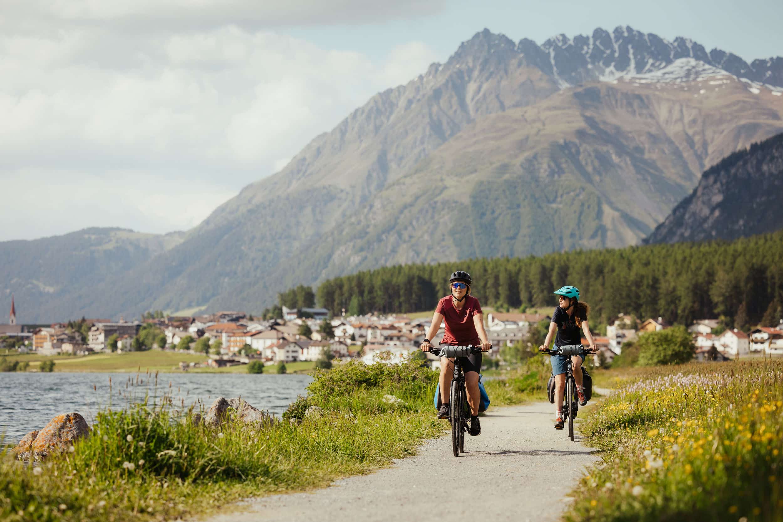 Zwei Radfahrer fahren am See entlang vor Bergkulisse und Dorf
