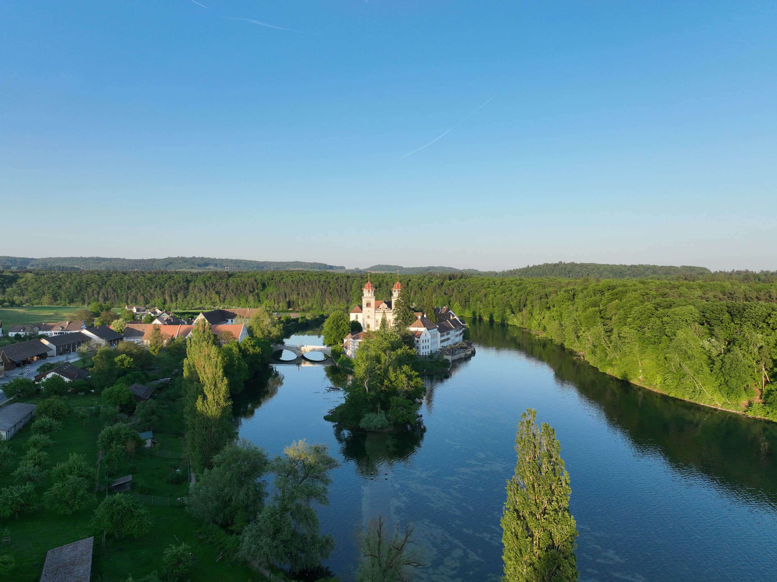 RheinWelten – Eine Reise entlang des Wassers © ProWeinland Fluss mit klosterartigem Gebäude auf einer Insel umgeben von Wald und Dorf