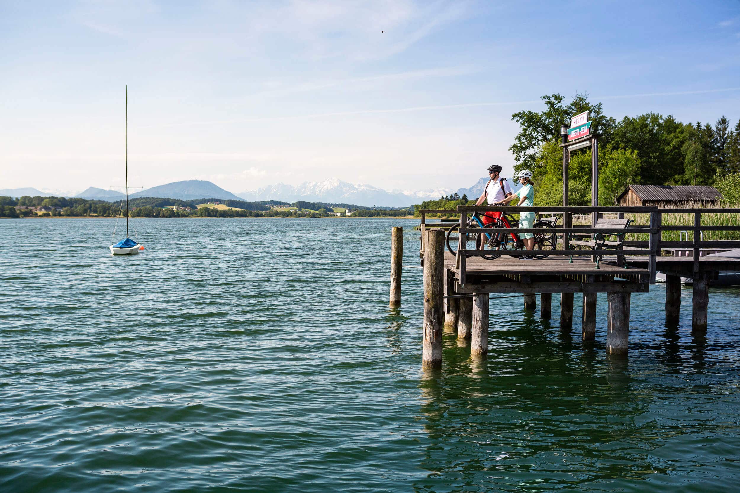 Zwei Radfahrer auf einem Steg am See mit Bergpanorama bei klarem Himmel