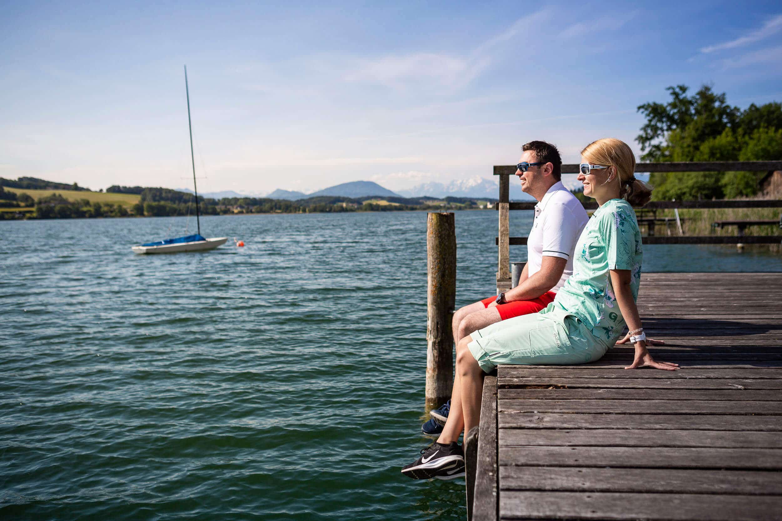 Paar sitzt auf einem Steg und blickt auf einen See mit Segelboot und Bergen