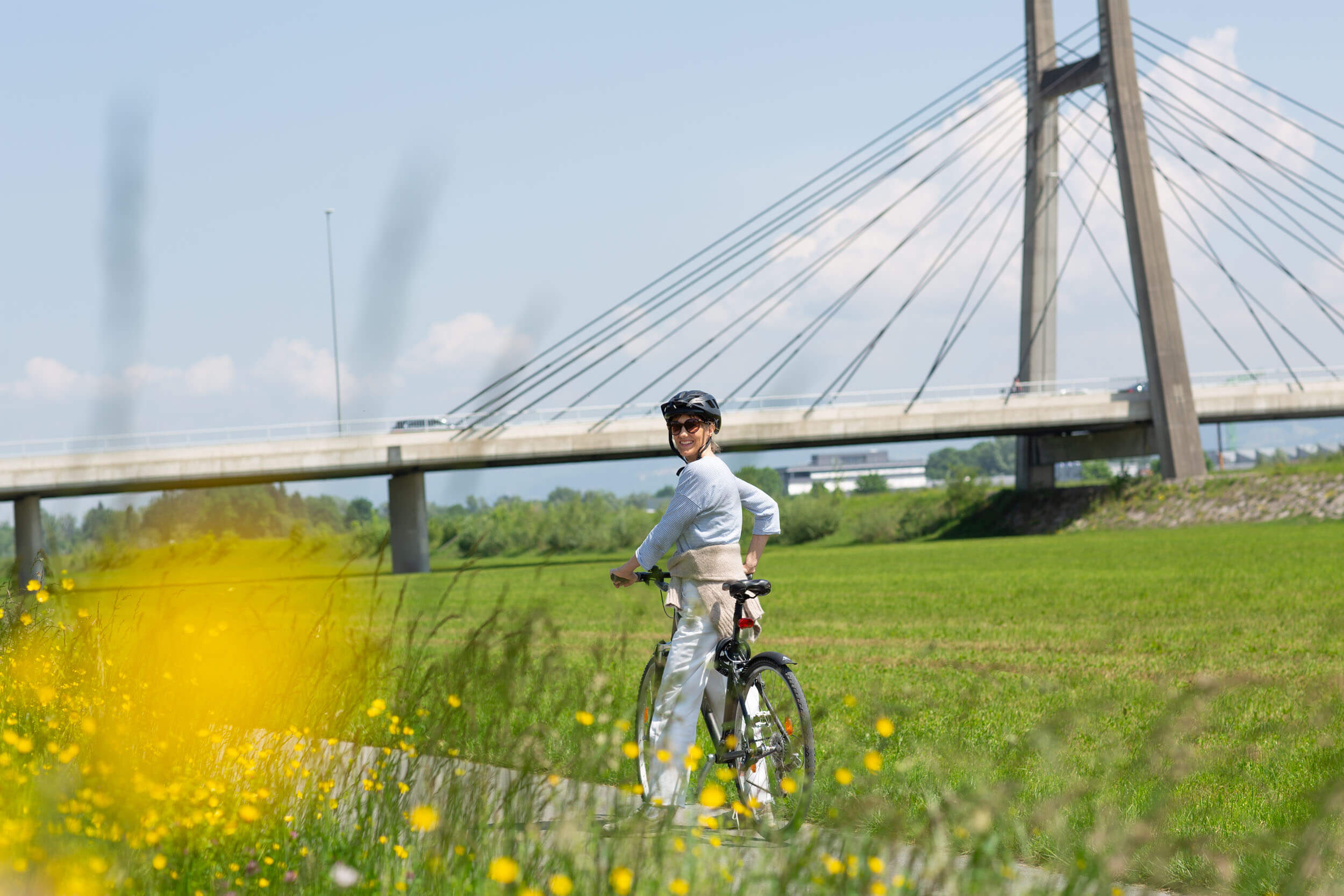 Frau mit Helm fährt Fahrrad vor einer Brücke in grüner Landschaft