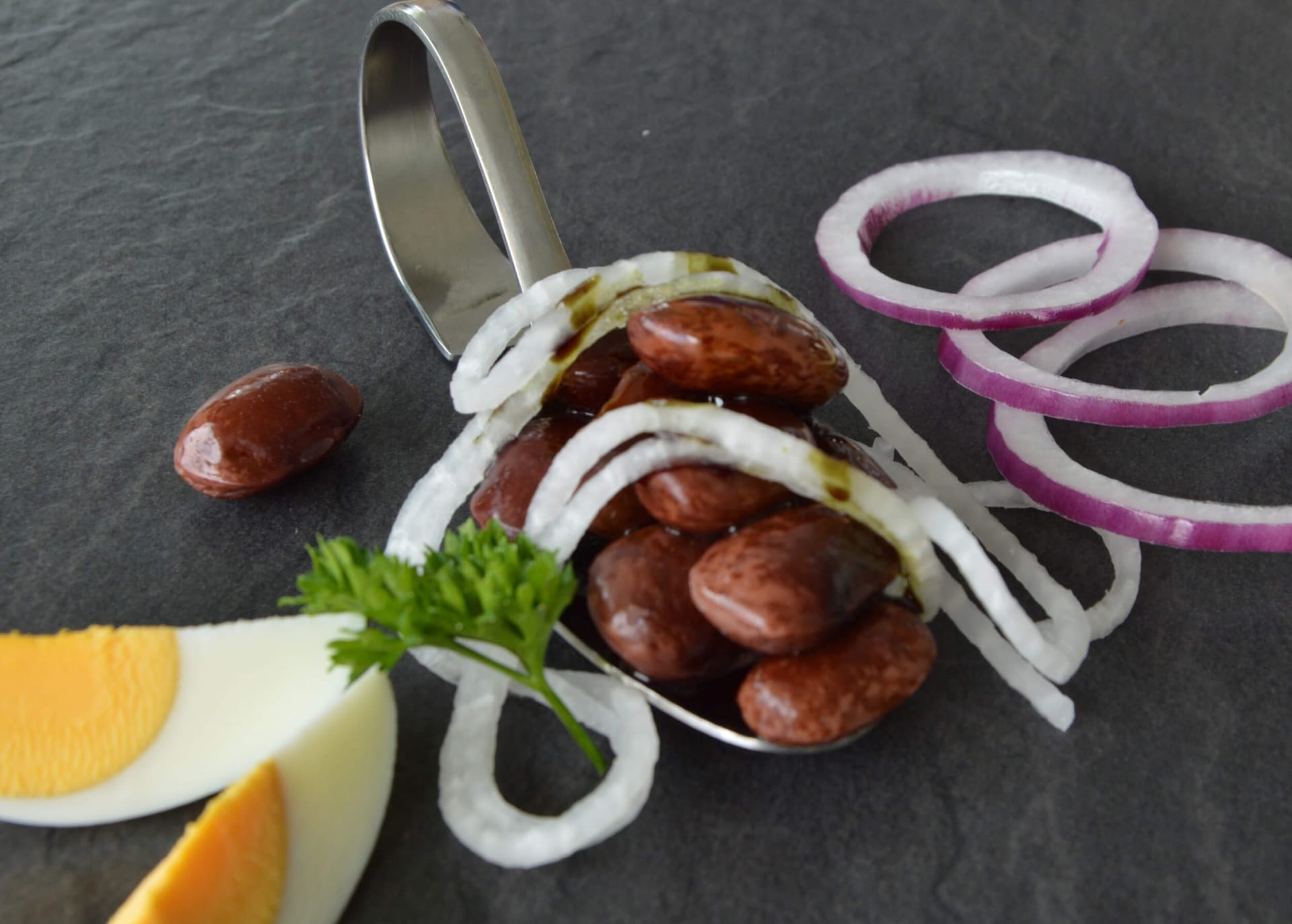 Spoon with red beans, onion rings, egg slices, and parsley on black surface